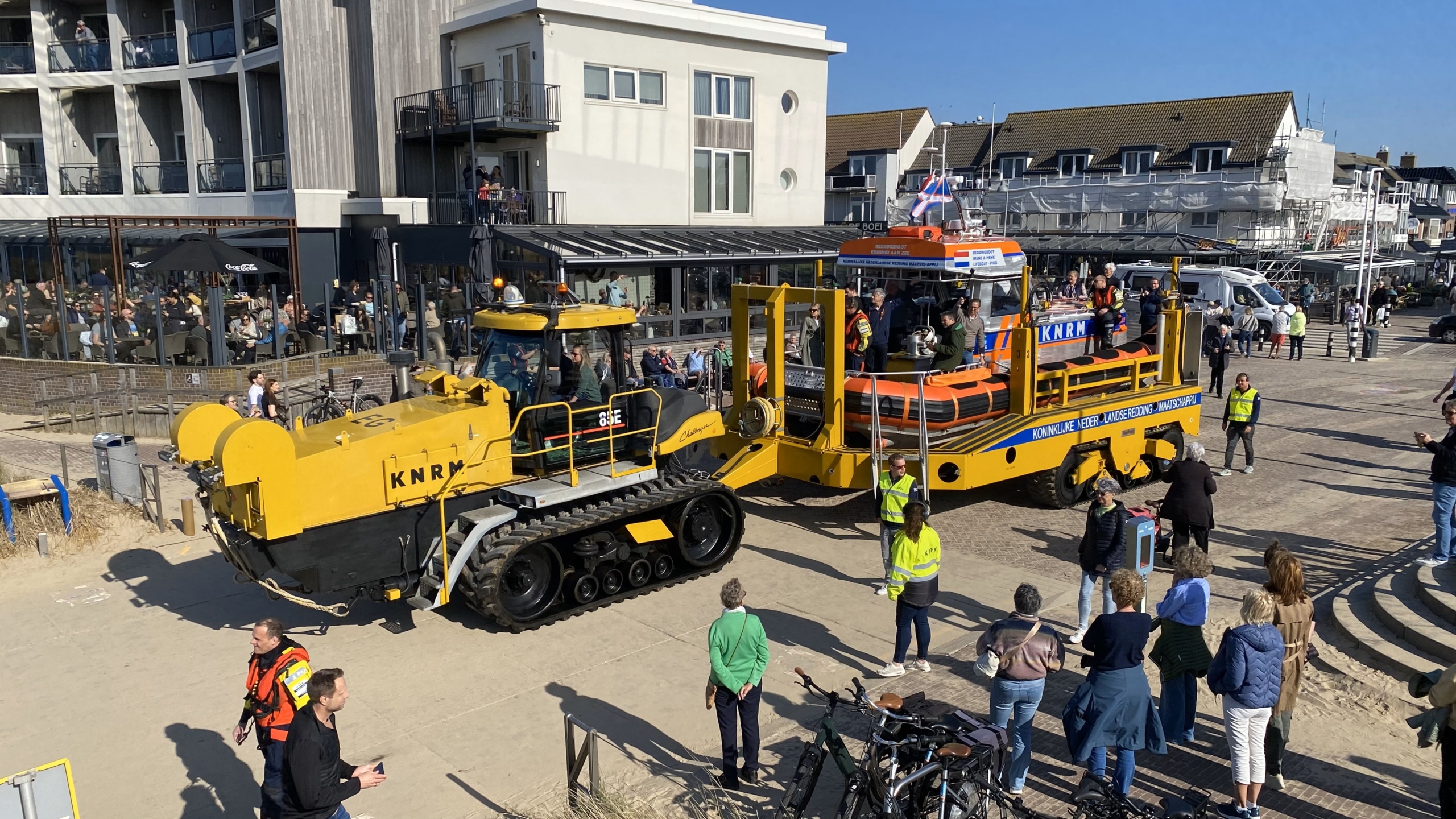 Geel voertuig van de KNRM dat een reddingsboot vervoert, omgeven door mensen op een plein bij een gebouw.