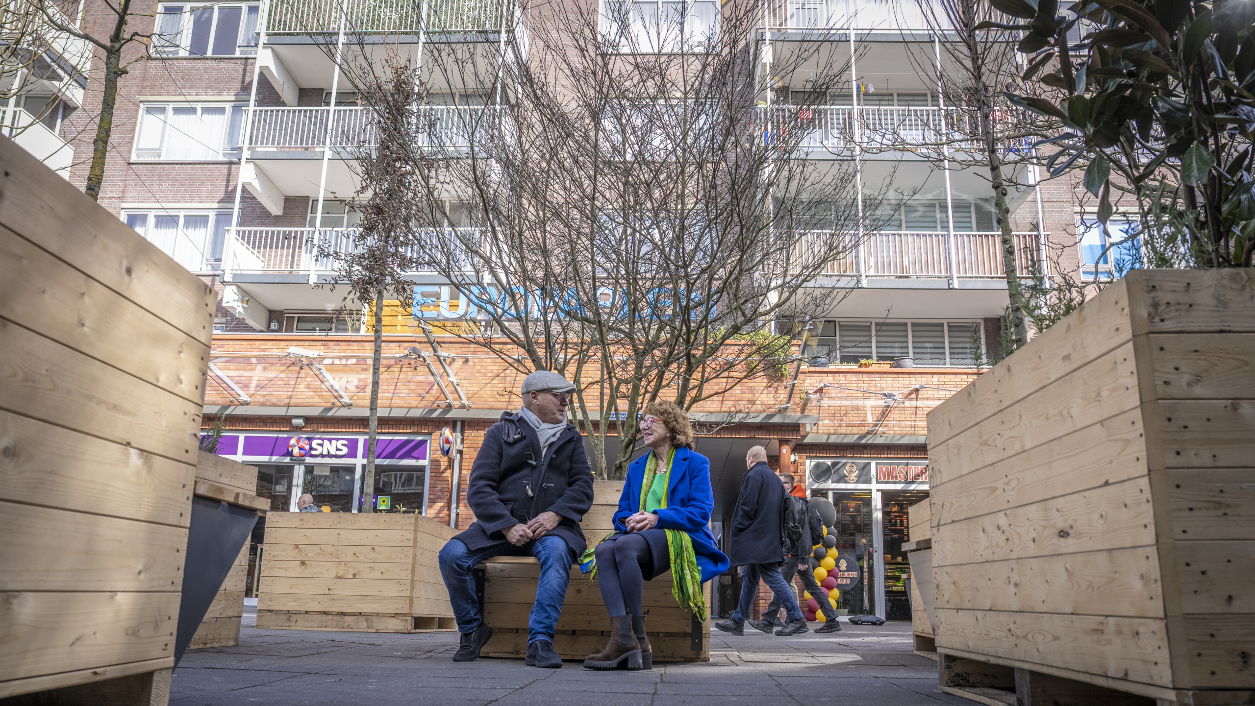 Twee mensen zittend op een bankje in een stedelijke omgeving met gebouwen en winkels op de achtergrond.