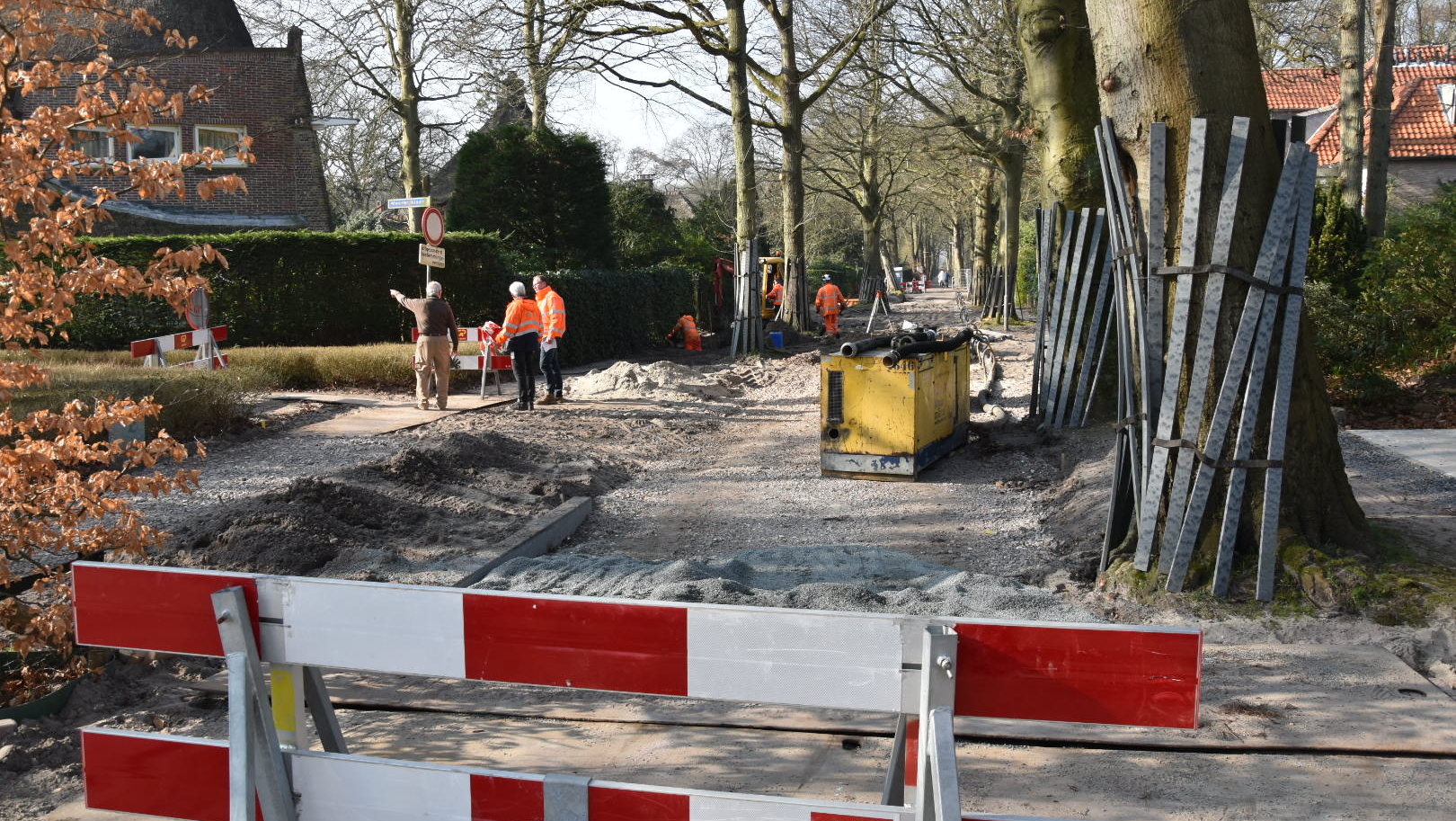 Wegwerkzaamheden op een met bomen omzoomde straat; werklieden in oranje kleding en gereedschap zichtbaar.