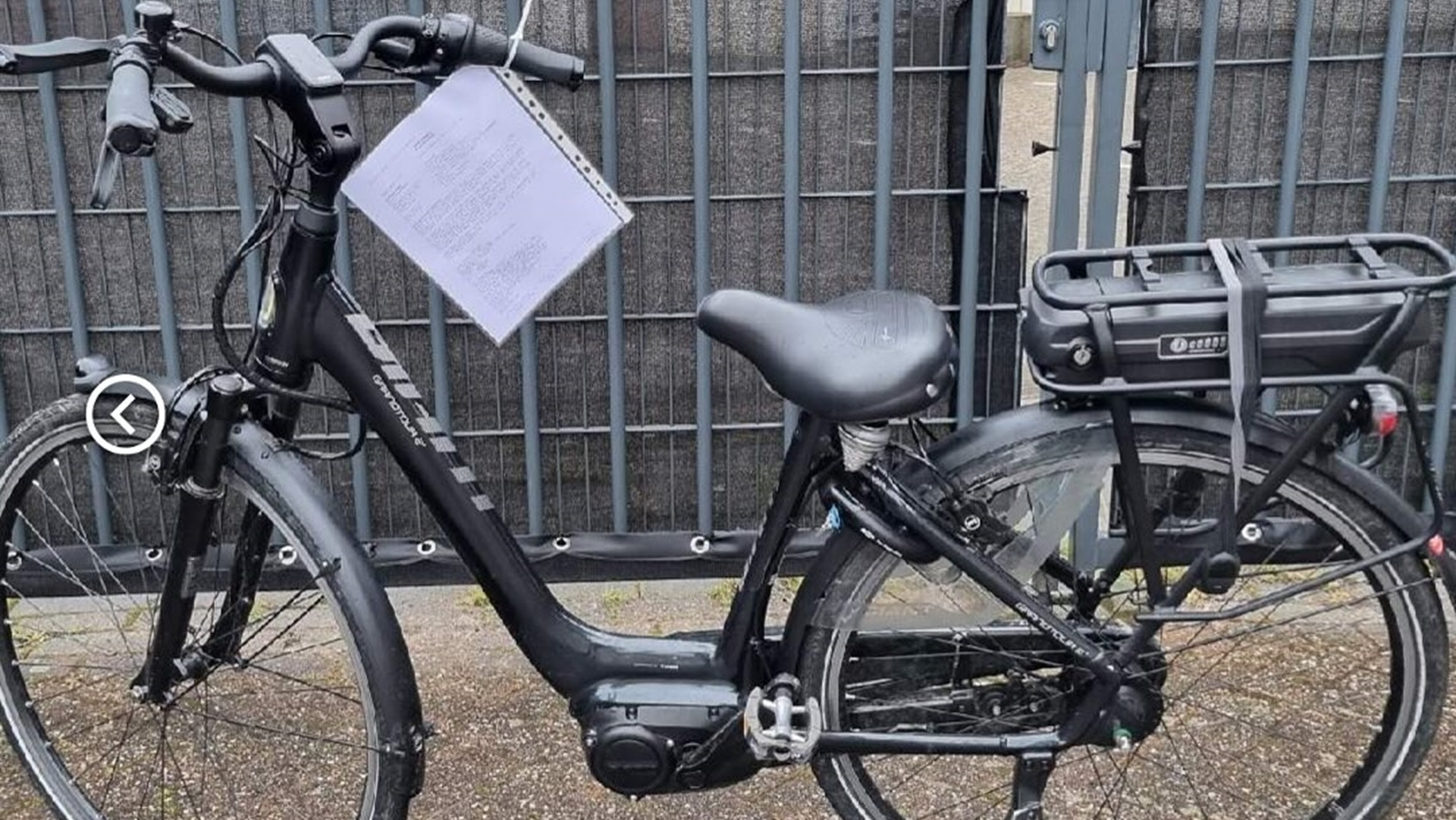 Zwarte elektrische fiets met een batterij op de bagagedrager, geparkeerd voor een hek met een briefje aan het stuur.