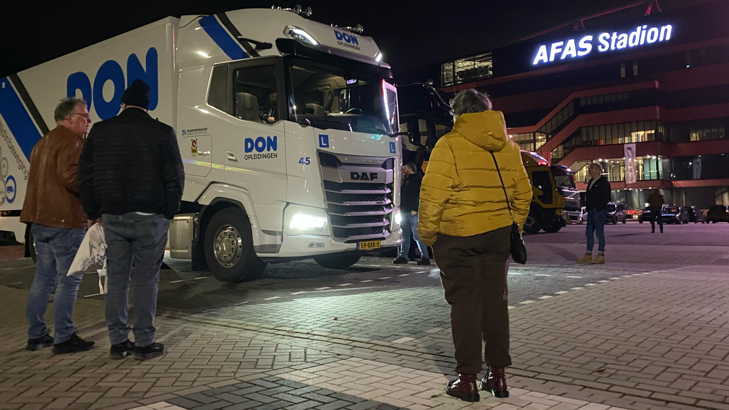 Mensen staan bij een vrachtwagen met de tekst "DON Opleidingen" op een parkeerplaats bij nacht, met het AFAS Stadion op de achtergrond.