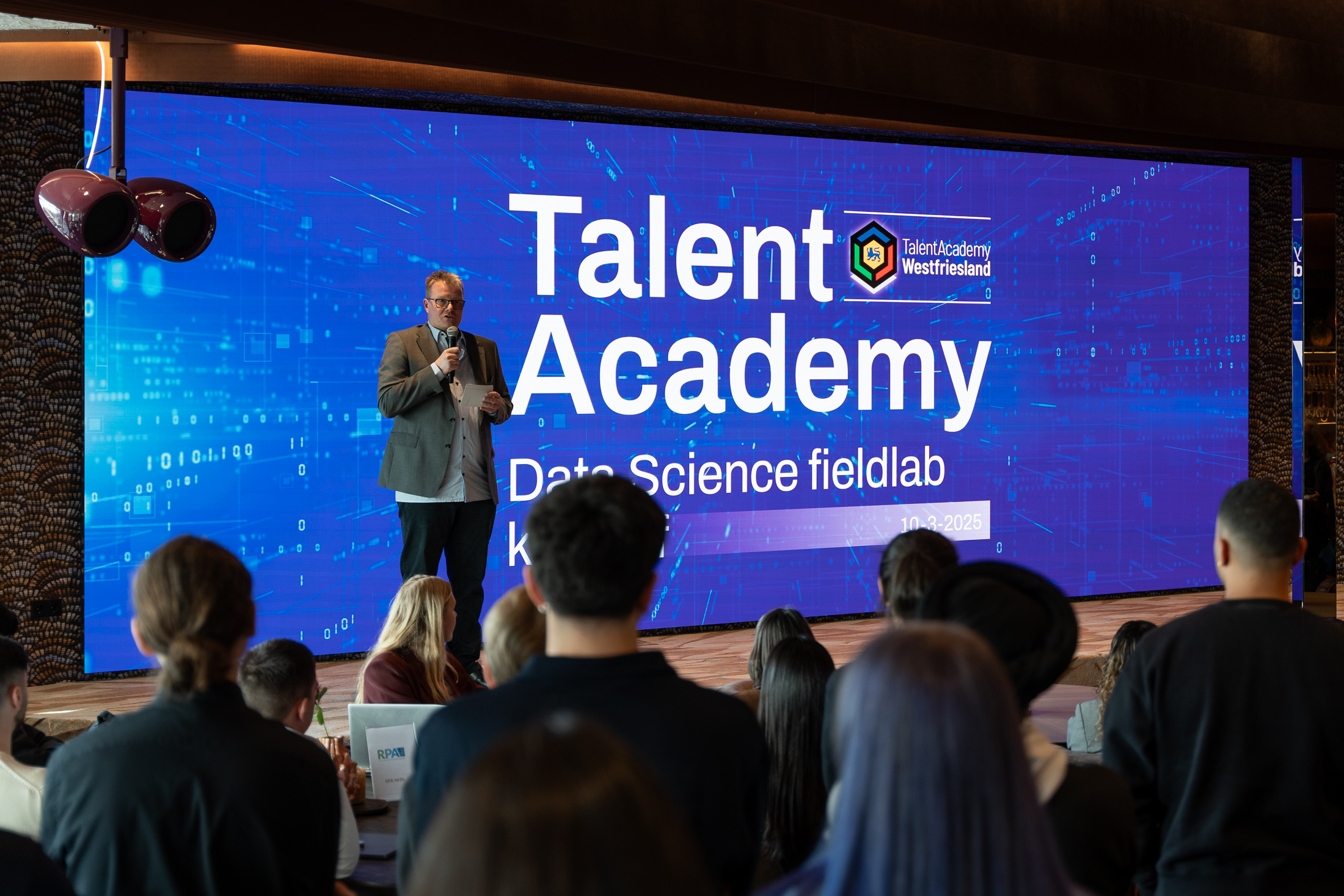 Man spreekt voor een groot scherm met "Talent Academy Data Science fieldlab" en publiek ervoor.