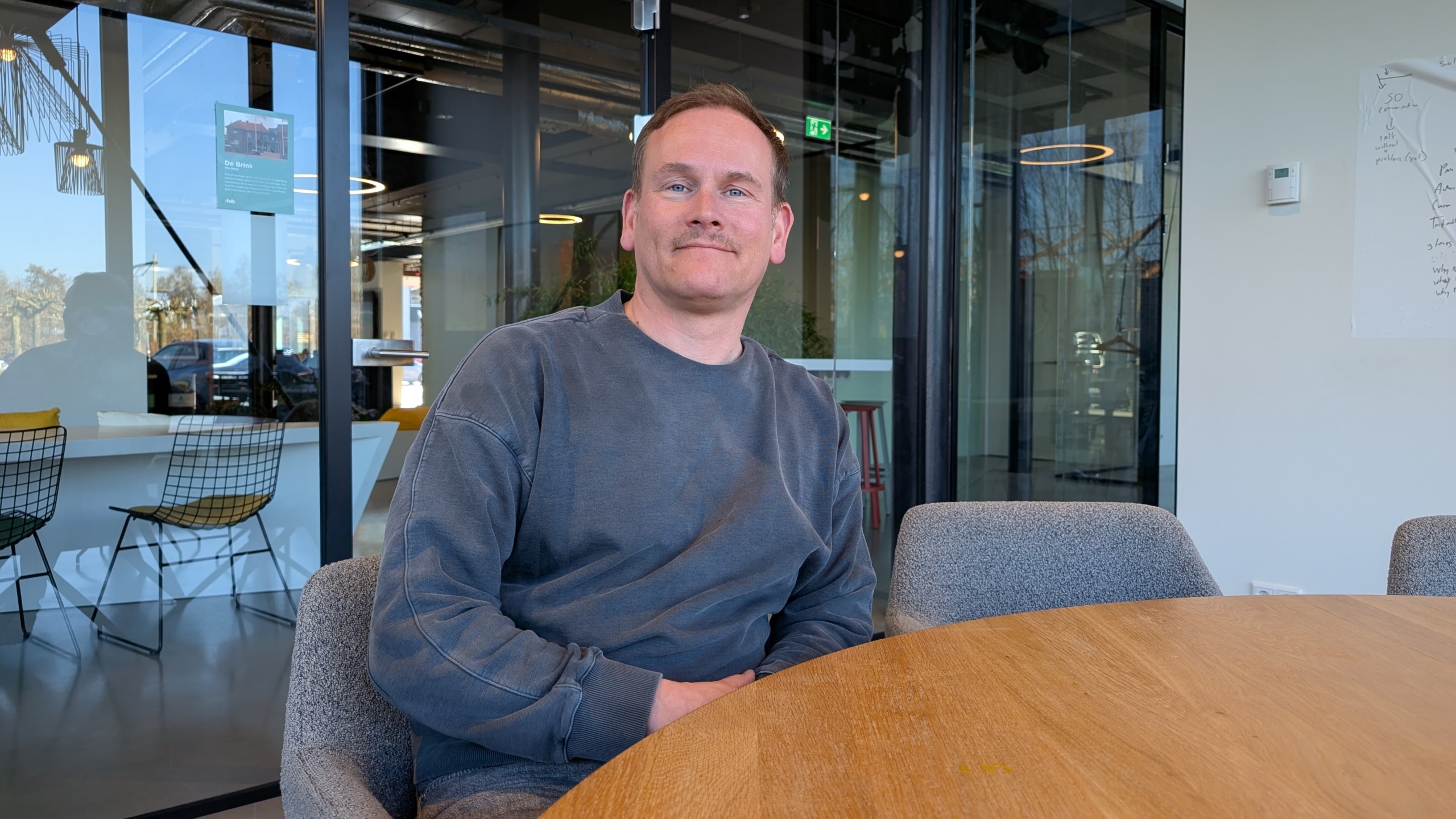 Een man zit aan een houten tafel in een moderne kantoorruimte met glazen wanden.