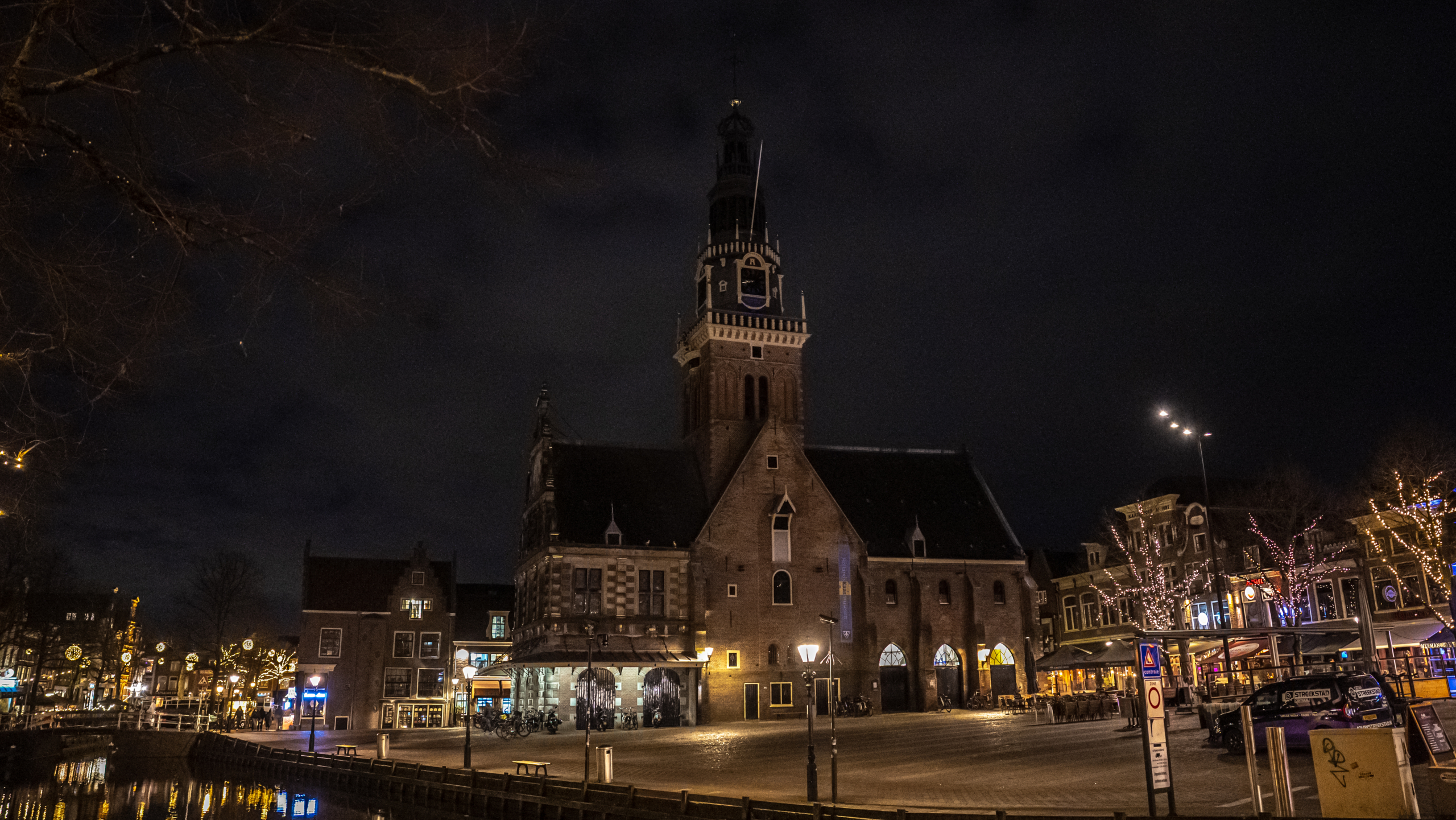 Een verlicht plein in de avond met een historisch gebouw, versierde bomen en panden met kerstverlichting.