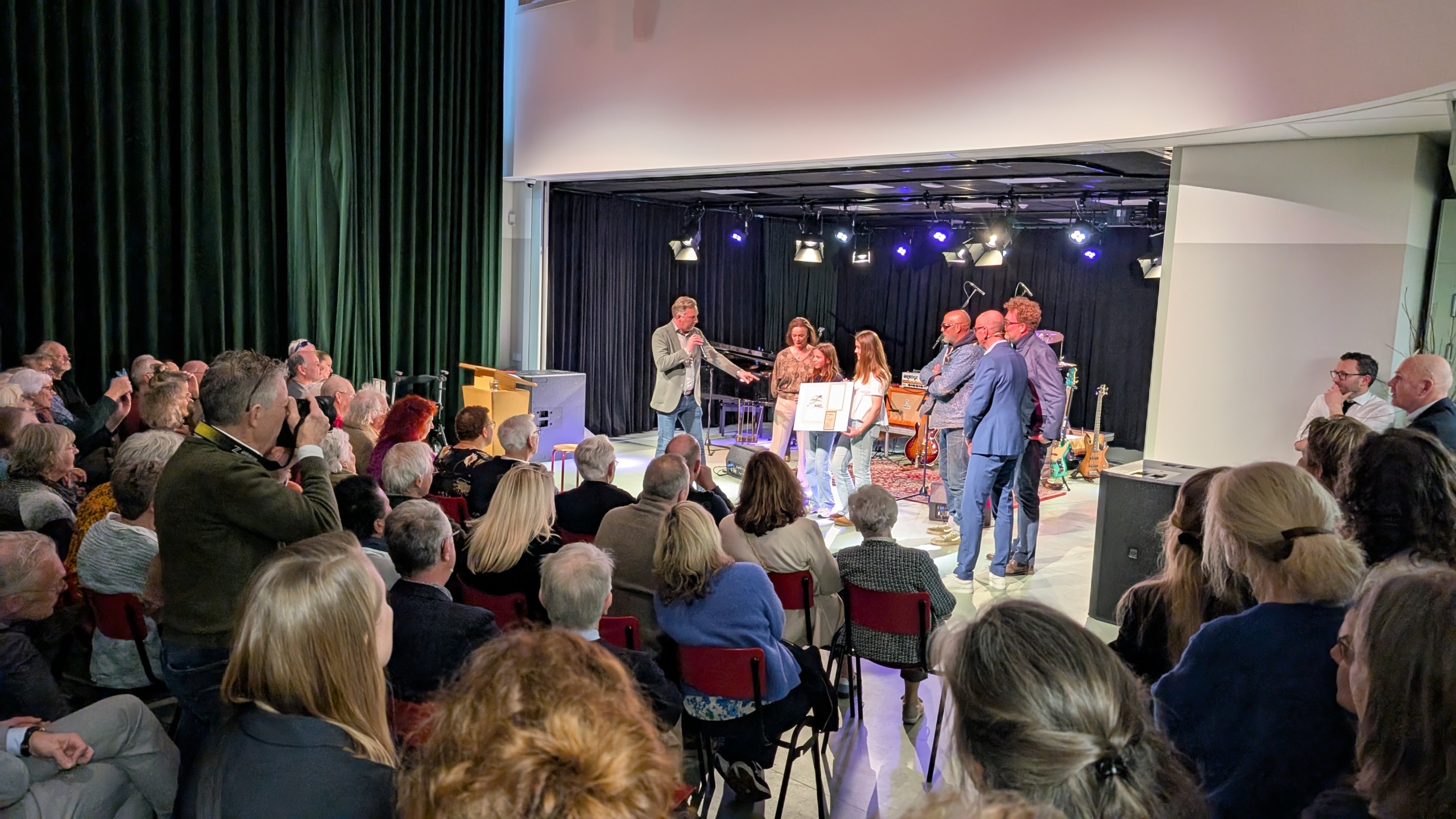 Mensengroep tijdens een evenement in een zaal met een spreker op een podium en een publiek dat luistert.