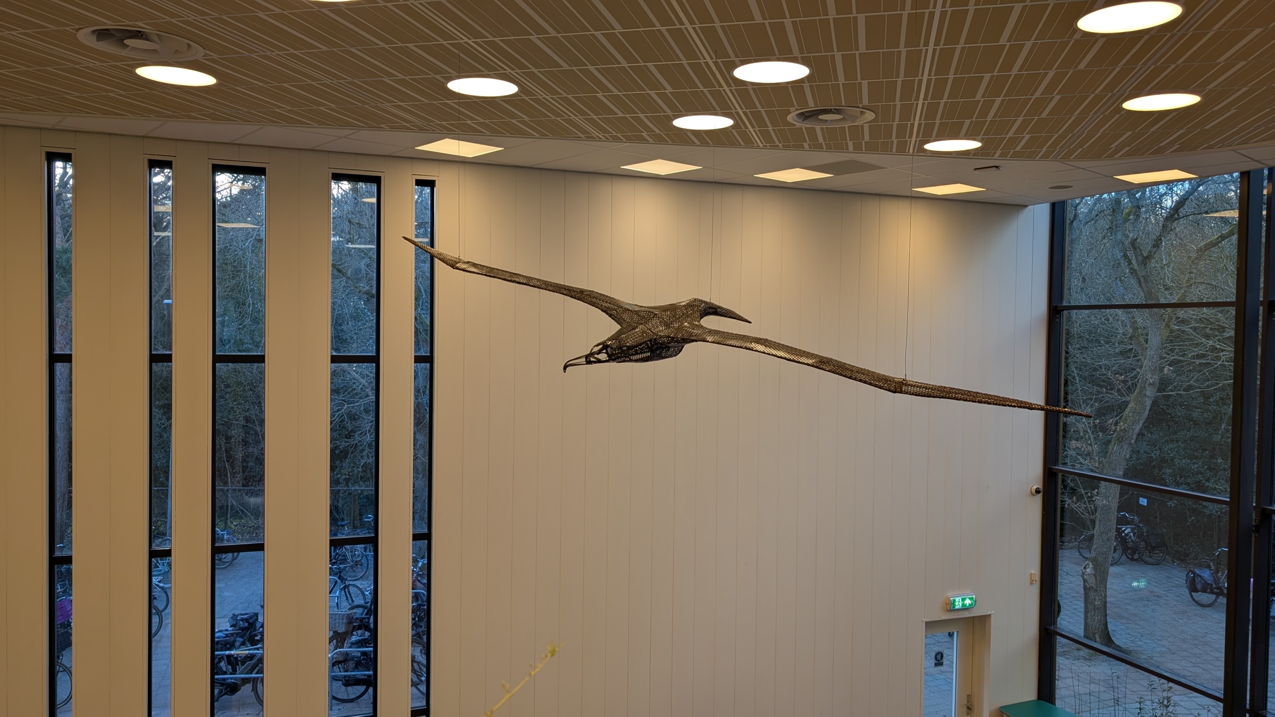 Hangende sculptuur van een vliegende vogel in een moderne ruimte met hoge ramen en plafondverlichting.