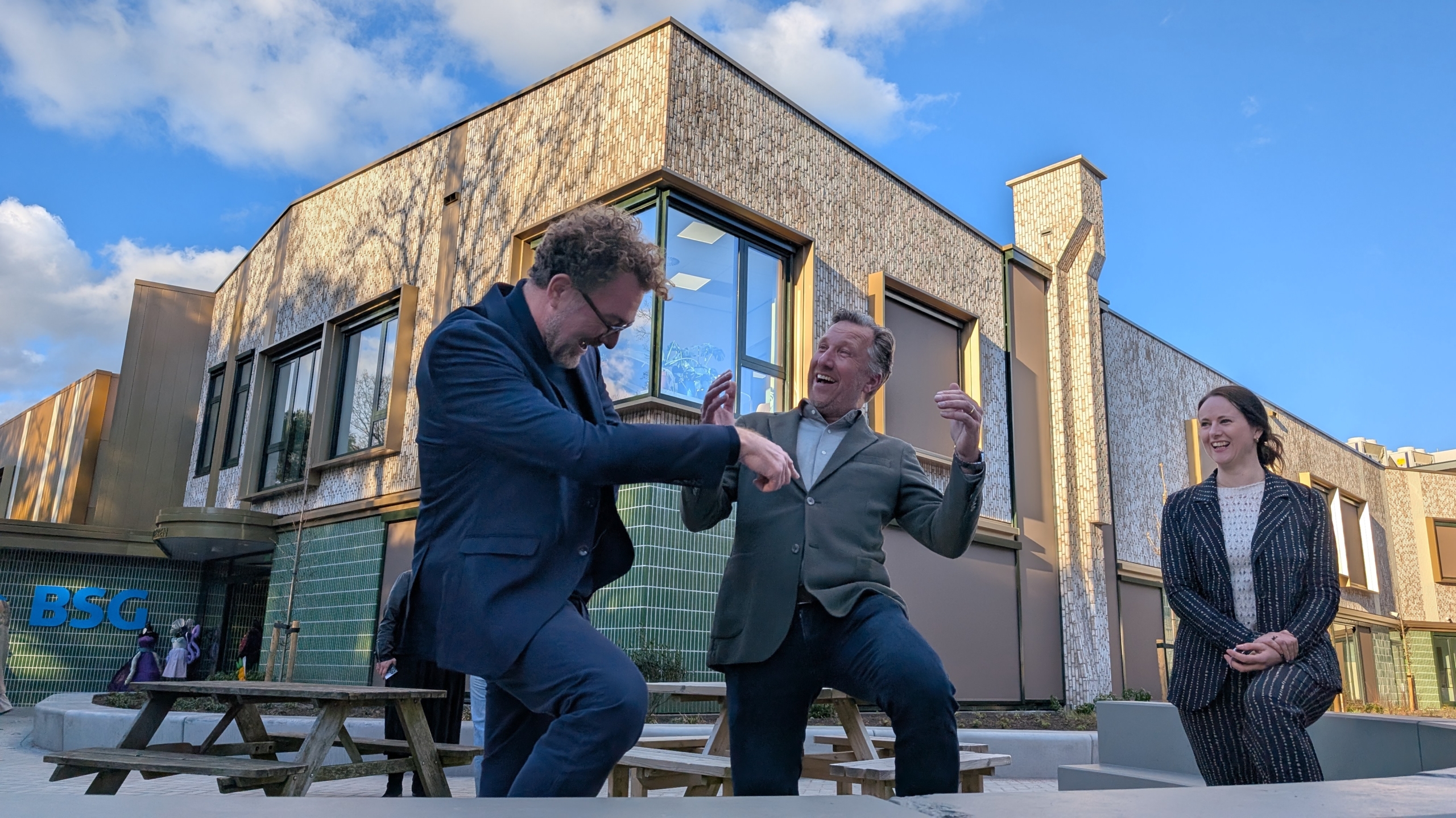 Twee mannen lachen en dansen samen voor een modern gebouw terwijl een vrouw glimlachend toekijkt.