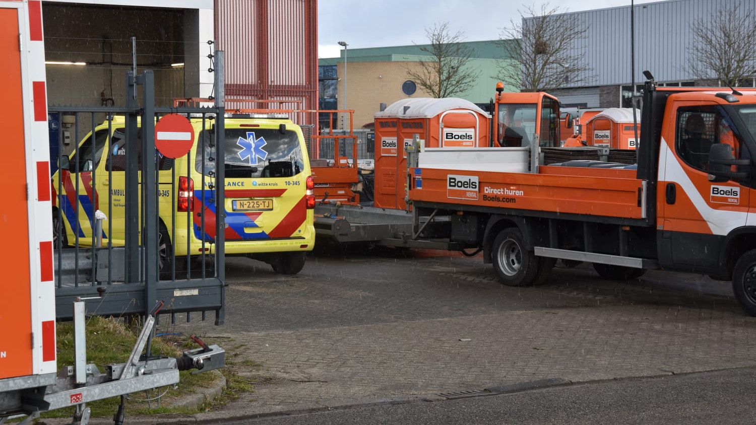 Ambulance en Boels vrachtwagen geparkeerd bij een gebouw, omgeven door oranje Boels apparatuur.