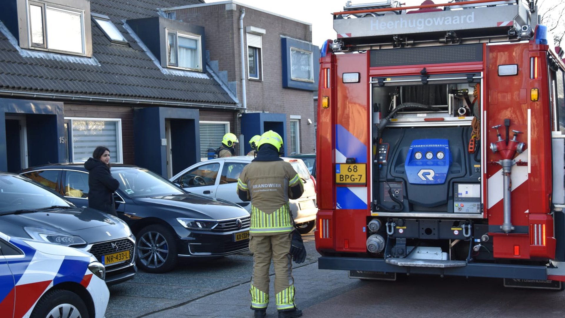 Brandweermannen in uniform staan naast een brandweerwagen voor een rijtjeshuis in een woonwijk, terwijl een vrouw in een zwarte jas toekijkt bij geparkeerde auto's.