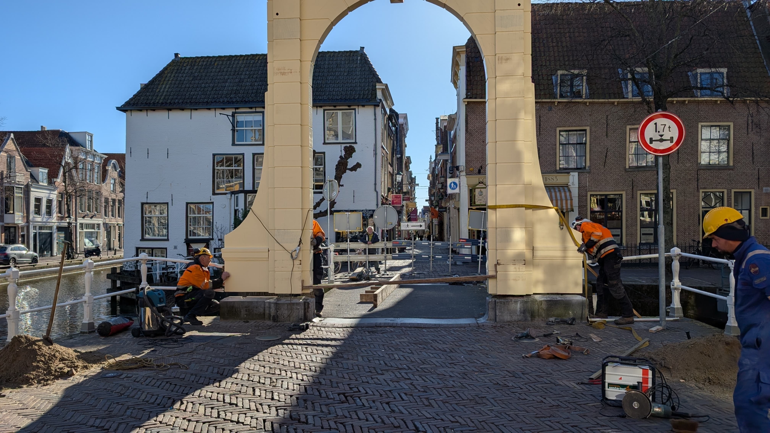 Werkzaamheden aan een brug met arbeiders in oranje veiligheidskleding en helmen bij een brugconstructie in een historisch stadscentrum met gebouwen en grachten.