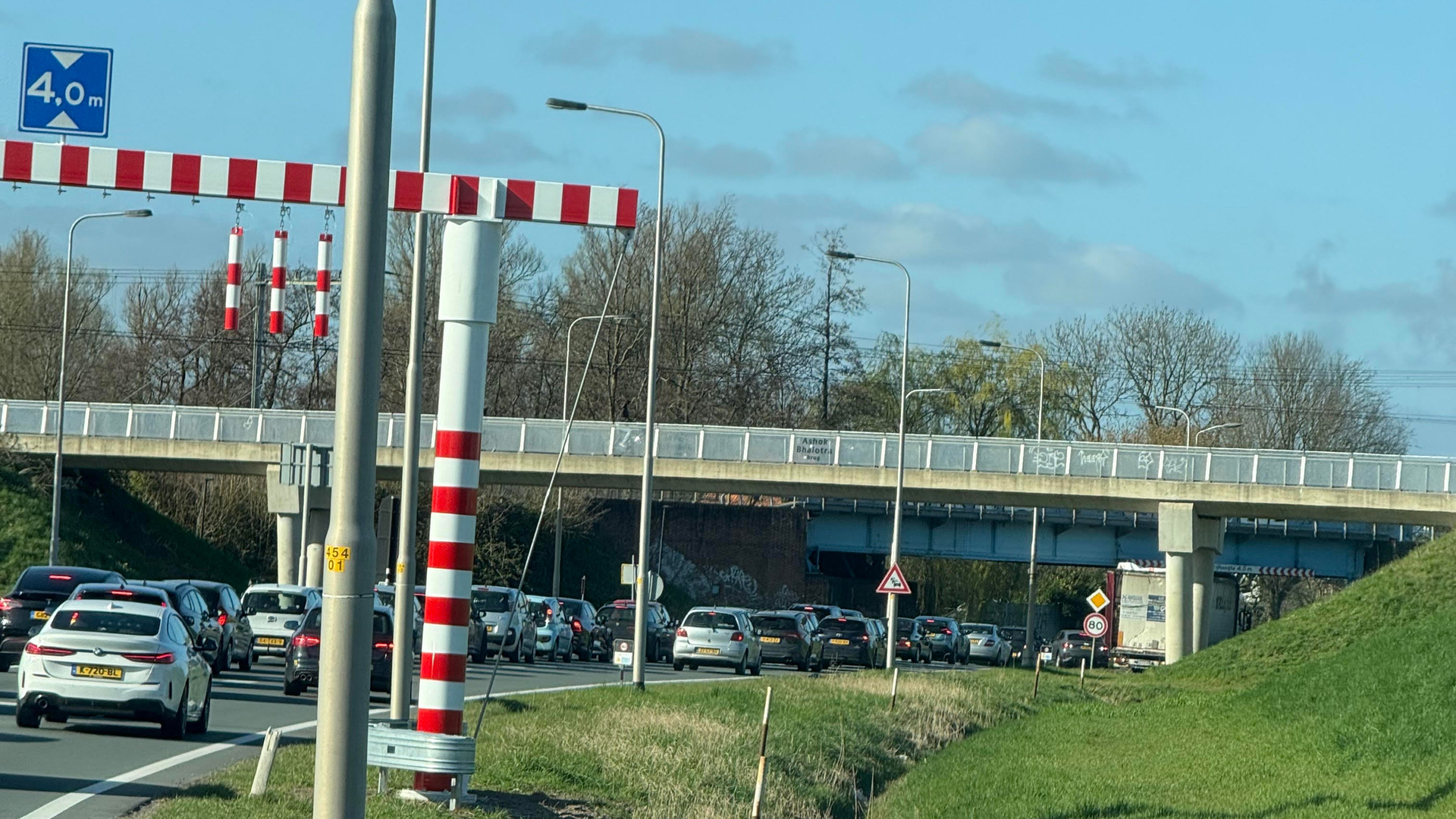 Verkeerstrook met een rij auto's die wachten onder een viaduct, met een hoogtebeperking van 4 meter aangegeven door rood-witte markeringen, onder een blauwe lucht.