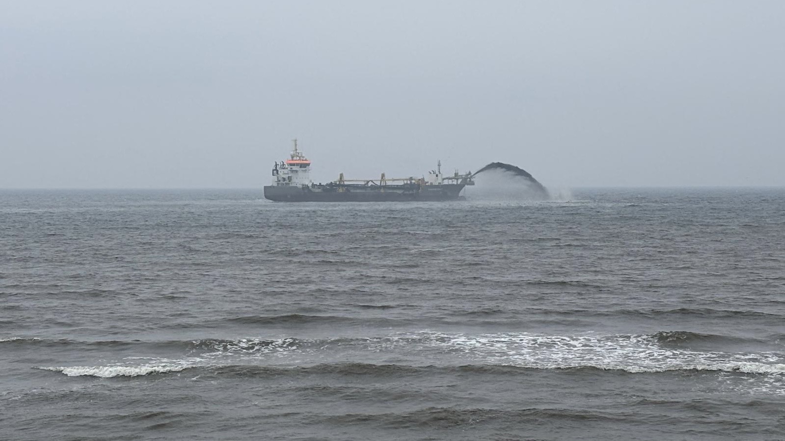 Een schip voert baggerwerkzaamheden uit op zee, waarbij een grote boog van materiaal in het water wordt gespoten, omgeven door een bewolkte lucht en kalme golven.