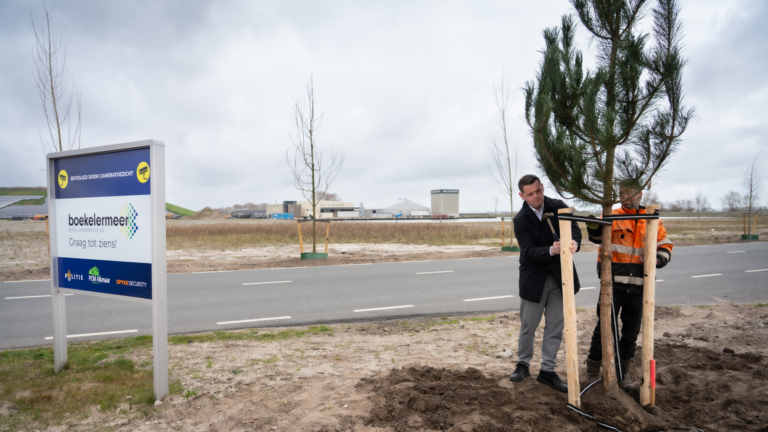 Twee mannen planten een boom naast een weg met een bord dat "boekelermeer" aangeeft, tegen een achtergrond van een industrieterrein met enkele gebouwen.