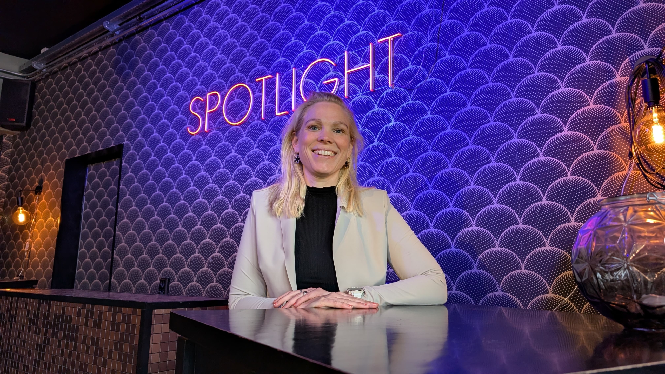 Een vrouw in een lichtgekleurde blazer en zwart shirt lacht terwijl ze voor een bar staat met een blauwe achtergrond en een neonbord met de tekst 'SPOTLIGHT'.