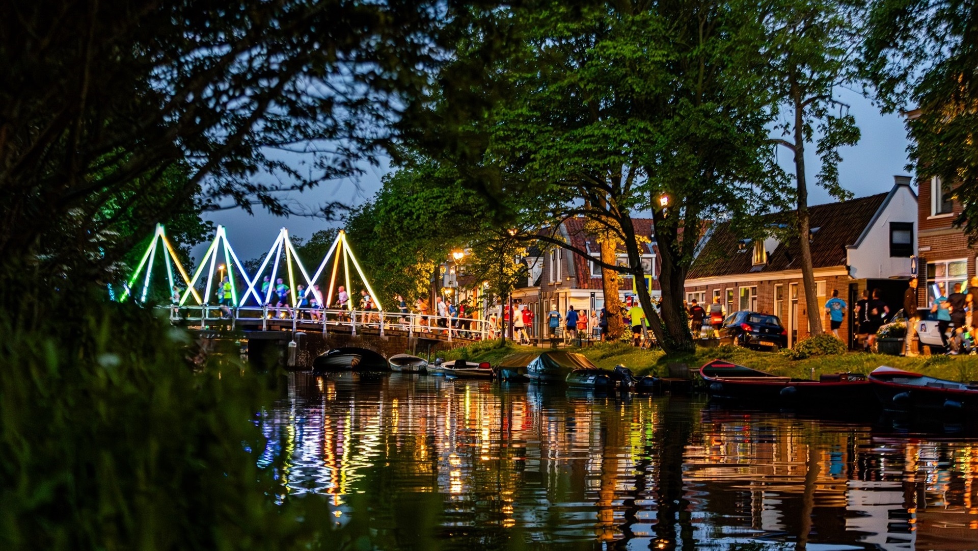 Verlichte voetgangersbrug met regenboogkleuren, omringd door bomen en huizen, weerspiegeld in een gracht.