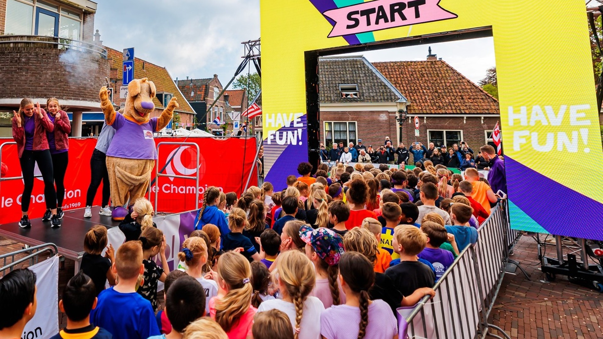 Kinderen verzamelen zich bij de startlijn van een evenement met een groot geel en paars 'Have Fun' bord en een persoon in een mascotte kostuum op een podium.