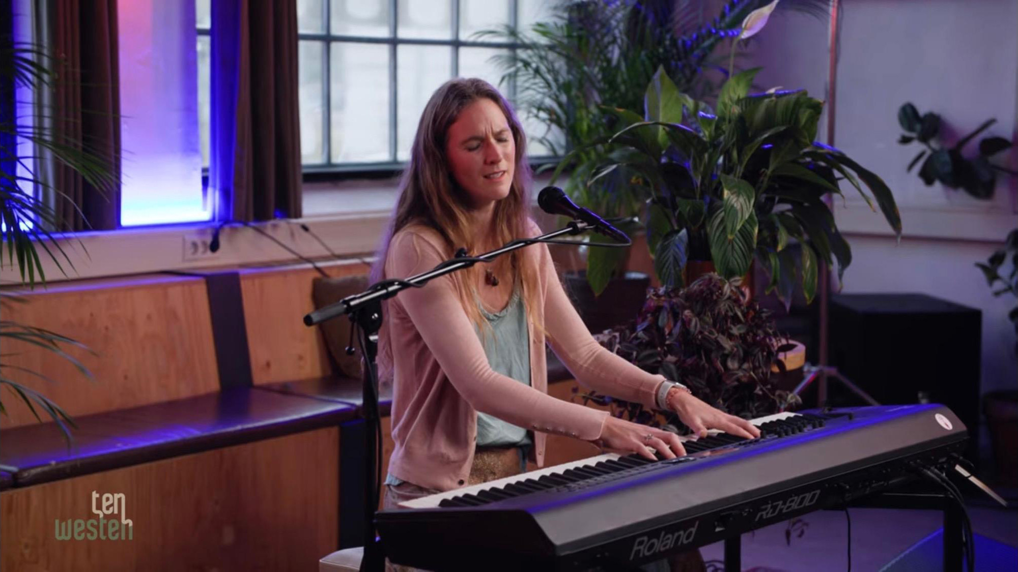 Vrouw speelt piano en zingt in een kamer met planten en kleurrijke verlichting.