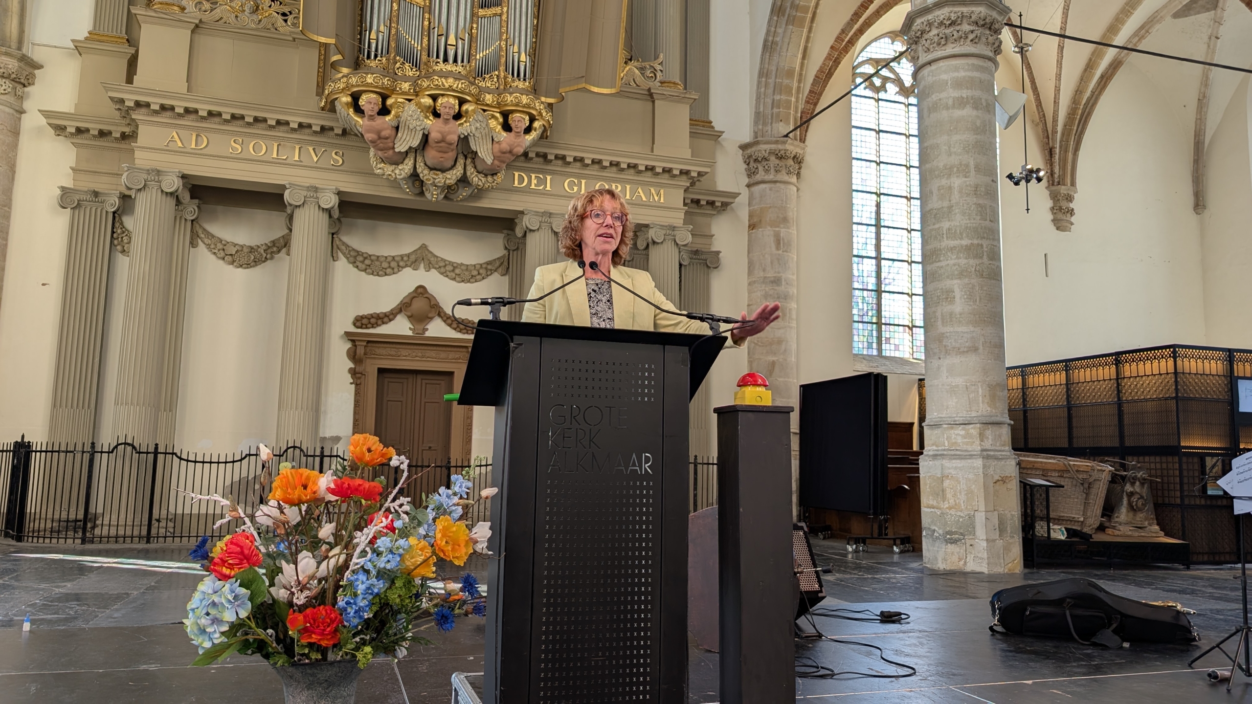 Een persoon staat achter een spreekgestoelte in de Grote Kerk van Alkmaar, omgeven door architectonische details en een boeket kleurrijke bloemen op de voorgrond.