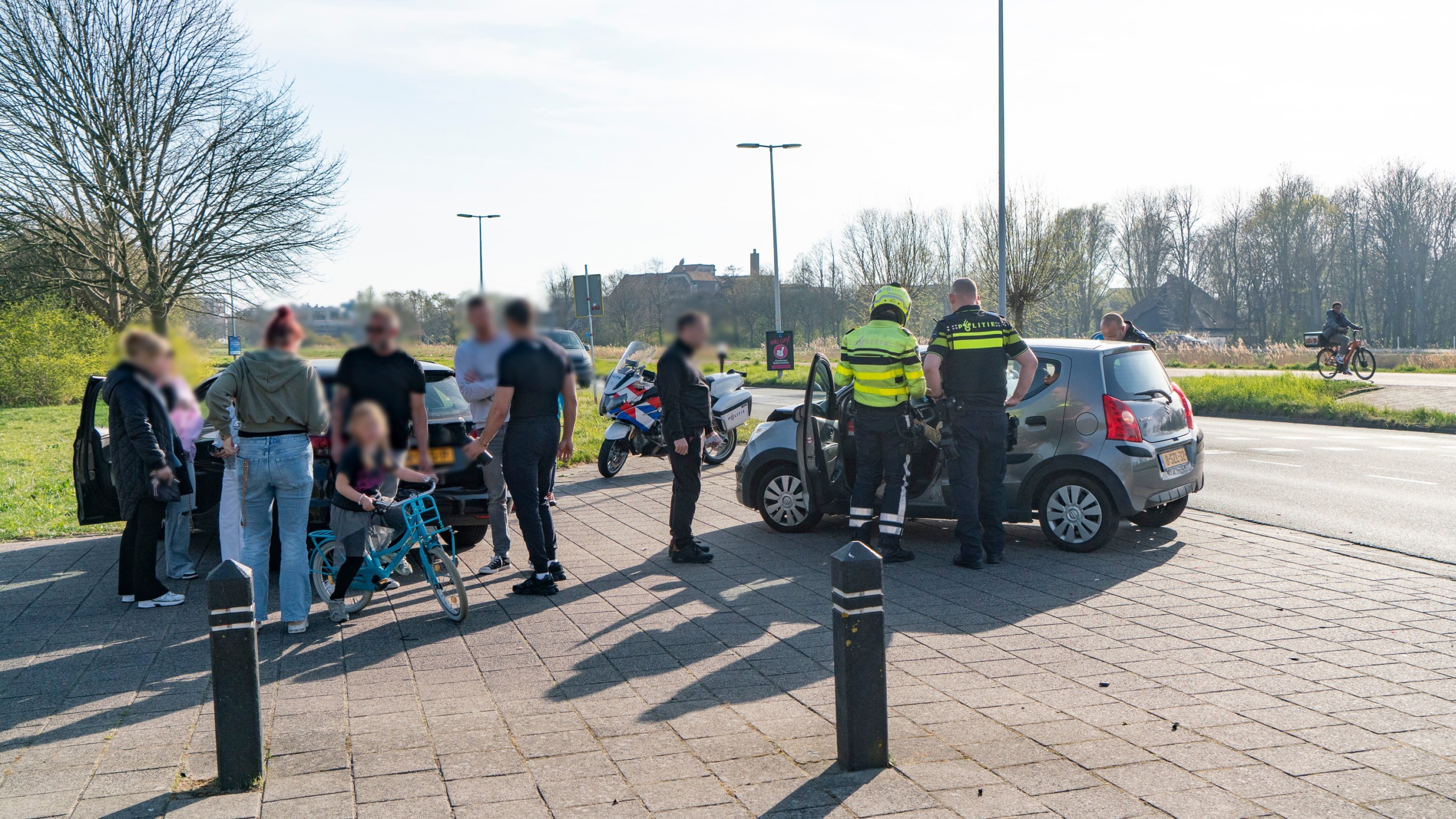 Groep mensen en politieagenten naast een auto bij een kruispunt; een motorfiets en twee fietsen zijn zichtbaar.