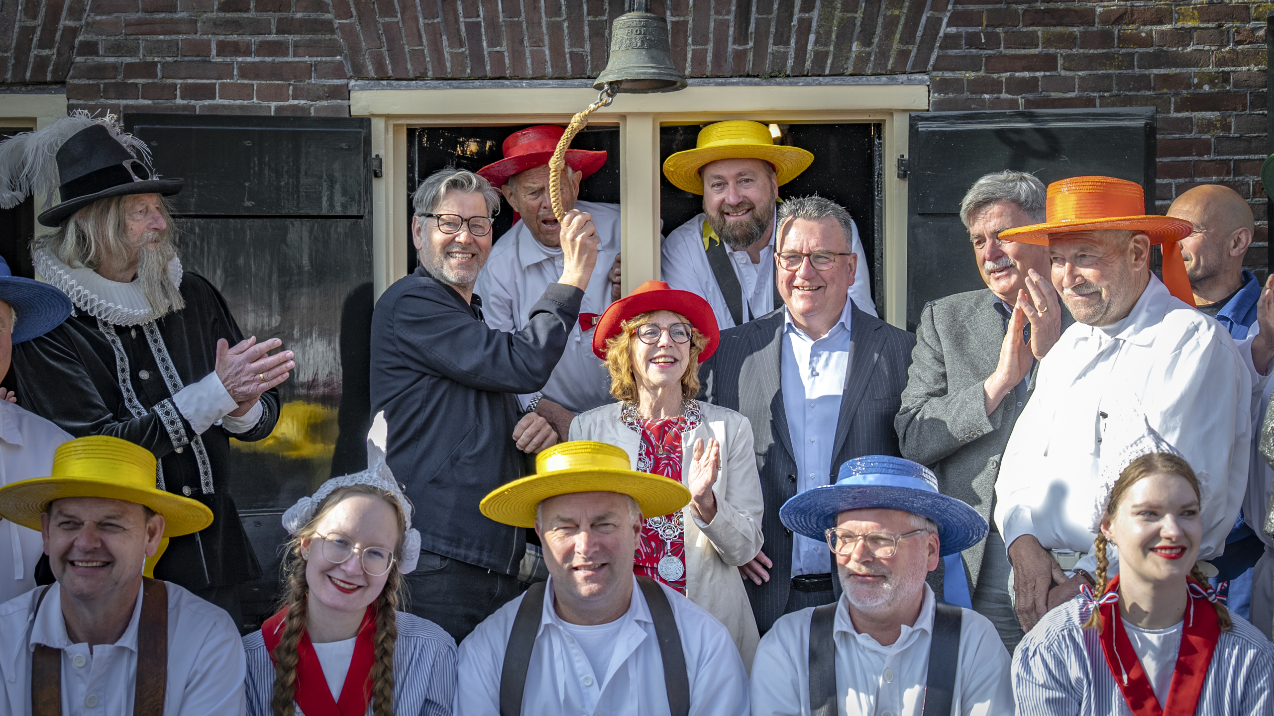 Groep mensen in traditionele klederdracht en moderne kleding, verzameld rond een bel met iemand die de bel luidt.