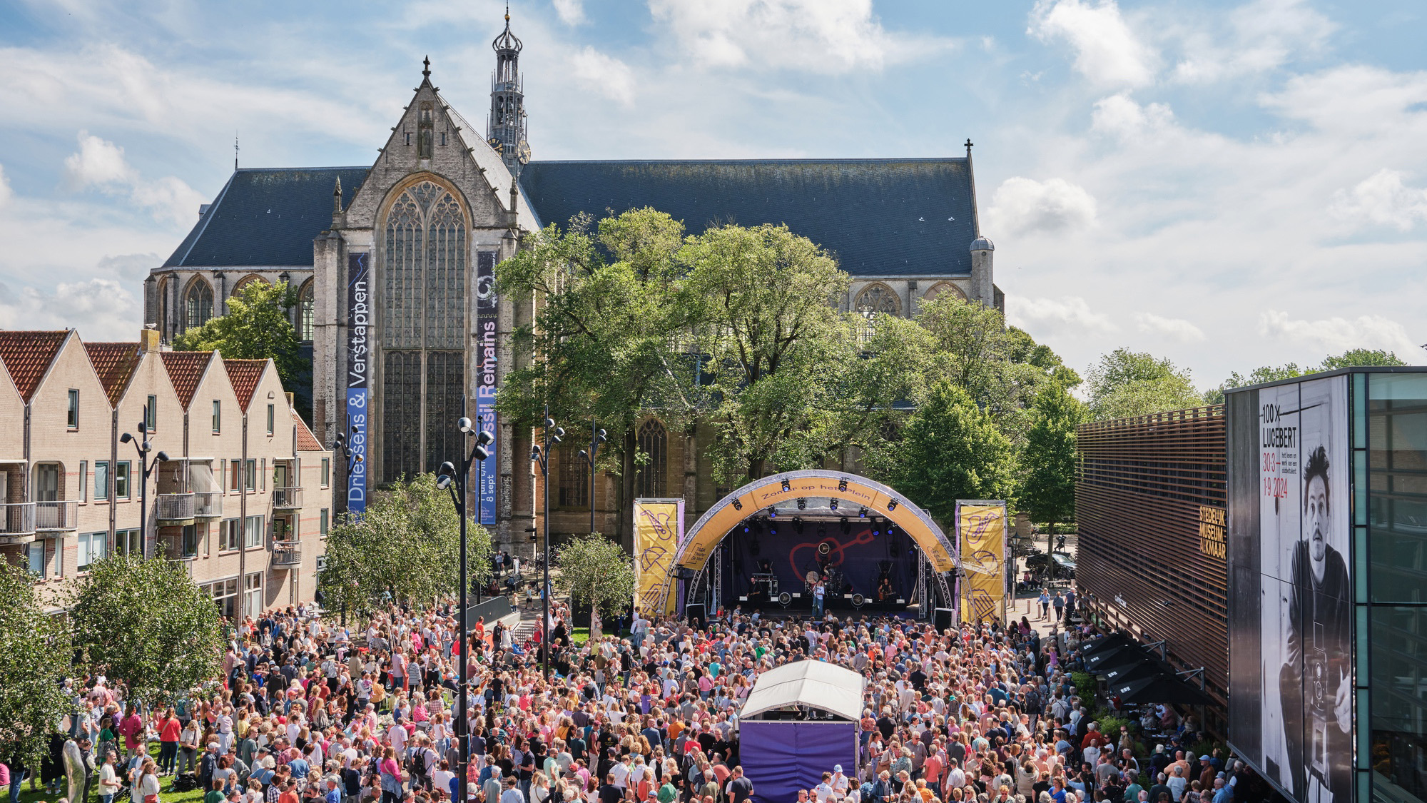 Een drukbezocht openluchtconcert voor de Grote Kerk van Breda, met een podium en een groot aantal toeschouwers op een zonnige dag.