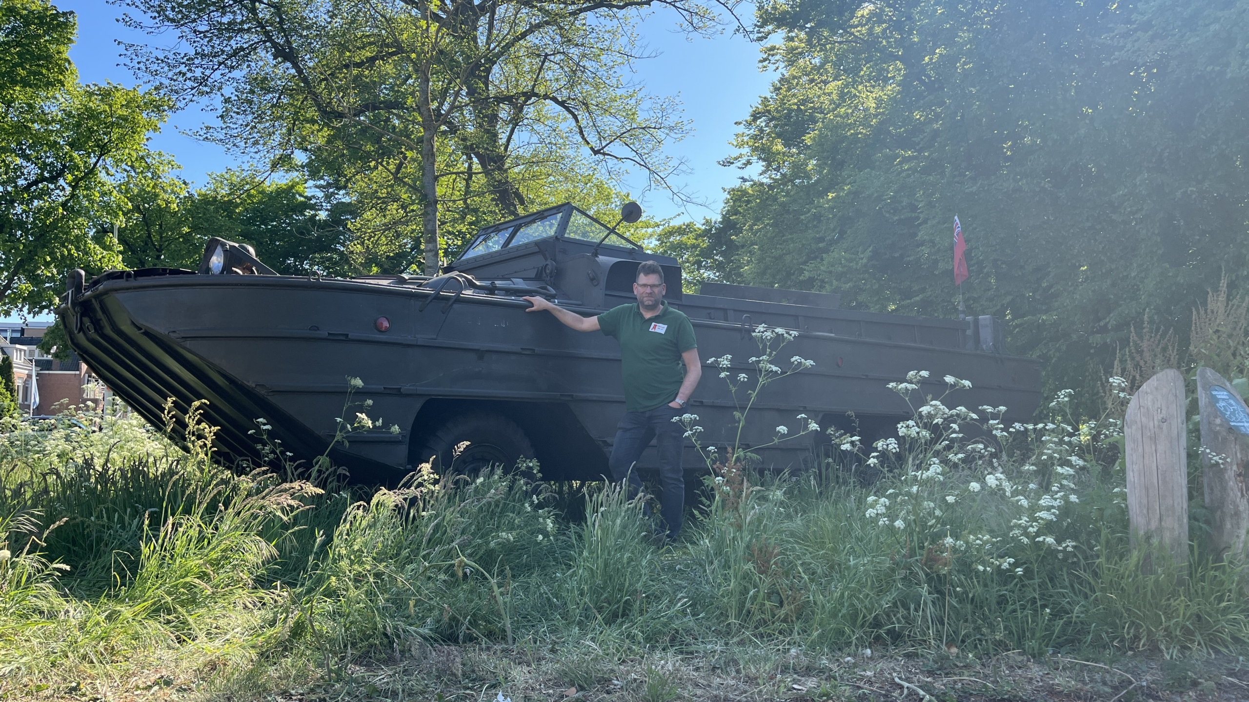 Man in groene trui leunt tegen een oude legervoertuig in een bosrijke omgeving, omgeven door gras en bloemen.