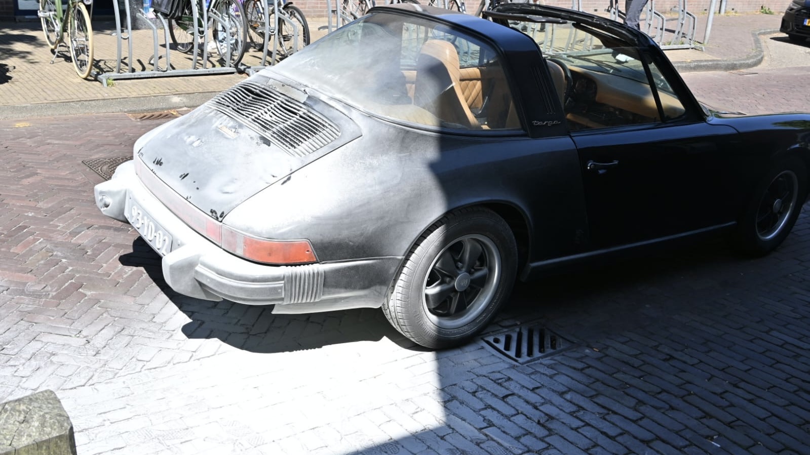Een zwarte klassieke sportauto, een Porsche Targa, geparkeerd op een straat met een kasseien oppervlak. De auto heeft een matte afwerking en het is zonnig weer, waardoor er schaduwen op het voertuig vallen.