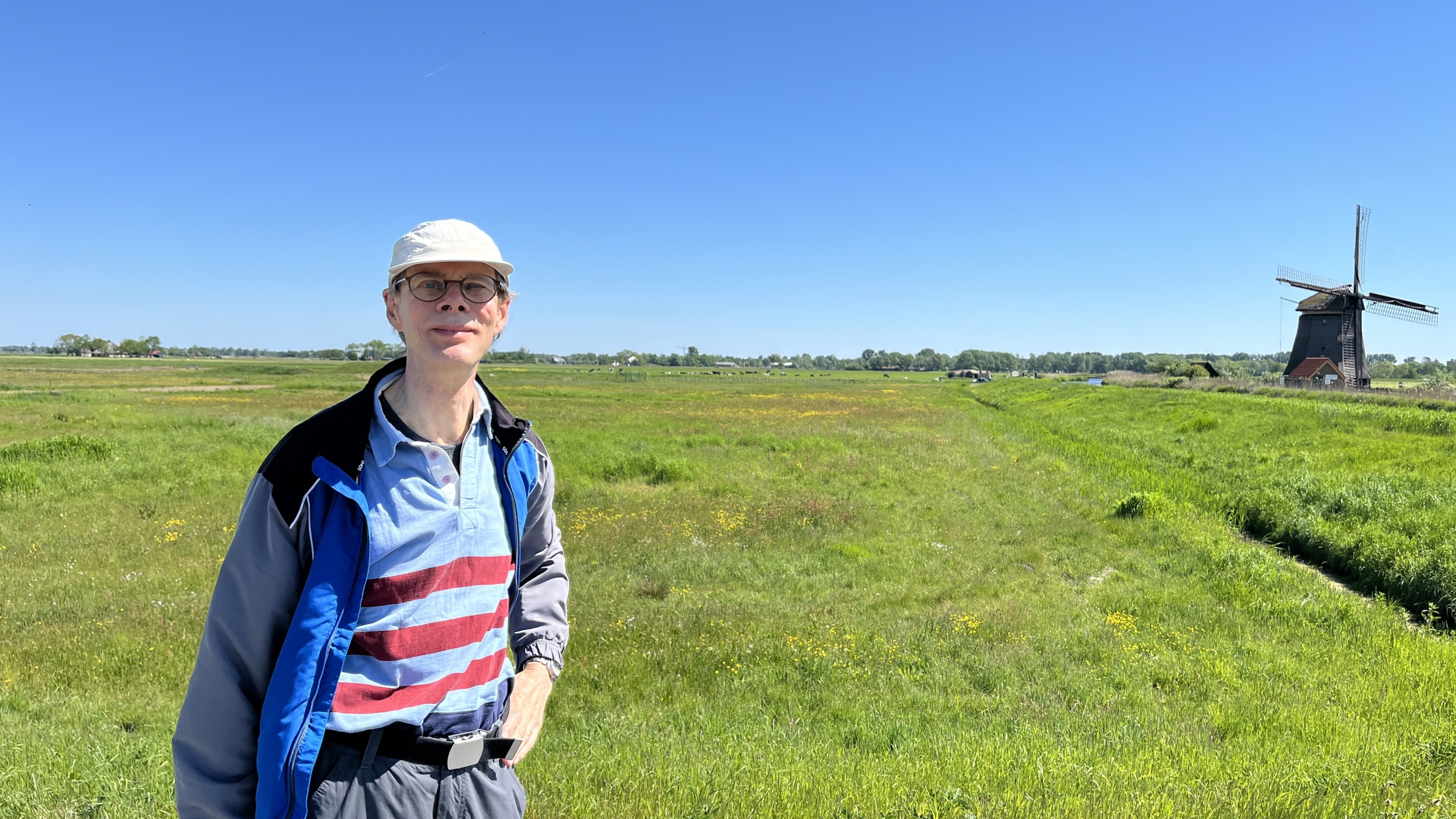 Man met een pet en bril staat in een groen weiland onder een heldere blauwe lucht, met een traditionele houten windmolen op de achtergrond.