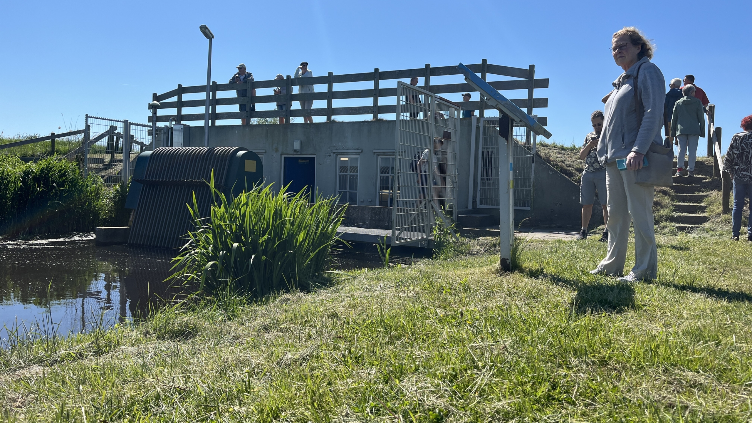Groep mensen staat buiten bij een gebouw met een hek en trap, omringd door gras en water, onder een heldere blauwe hemel.