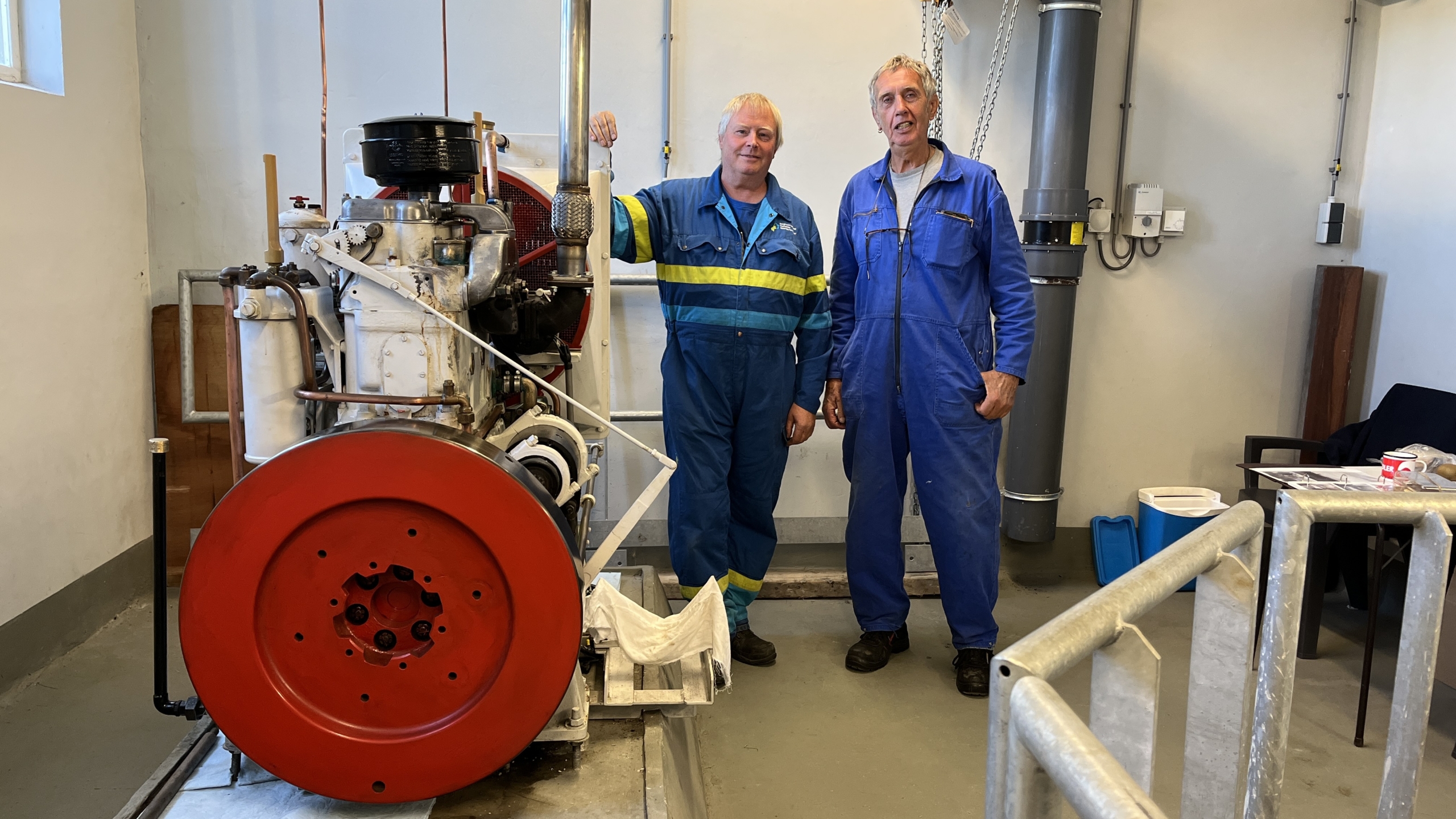 Twee mannen in blauwe overalls staan naast een grote, industriële machine met een rode schijf in een werkplaats.
