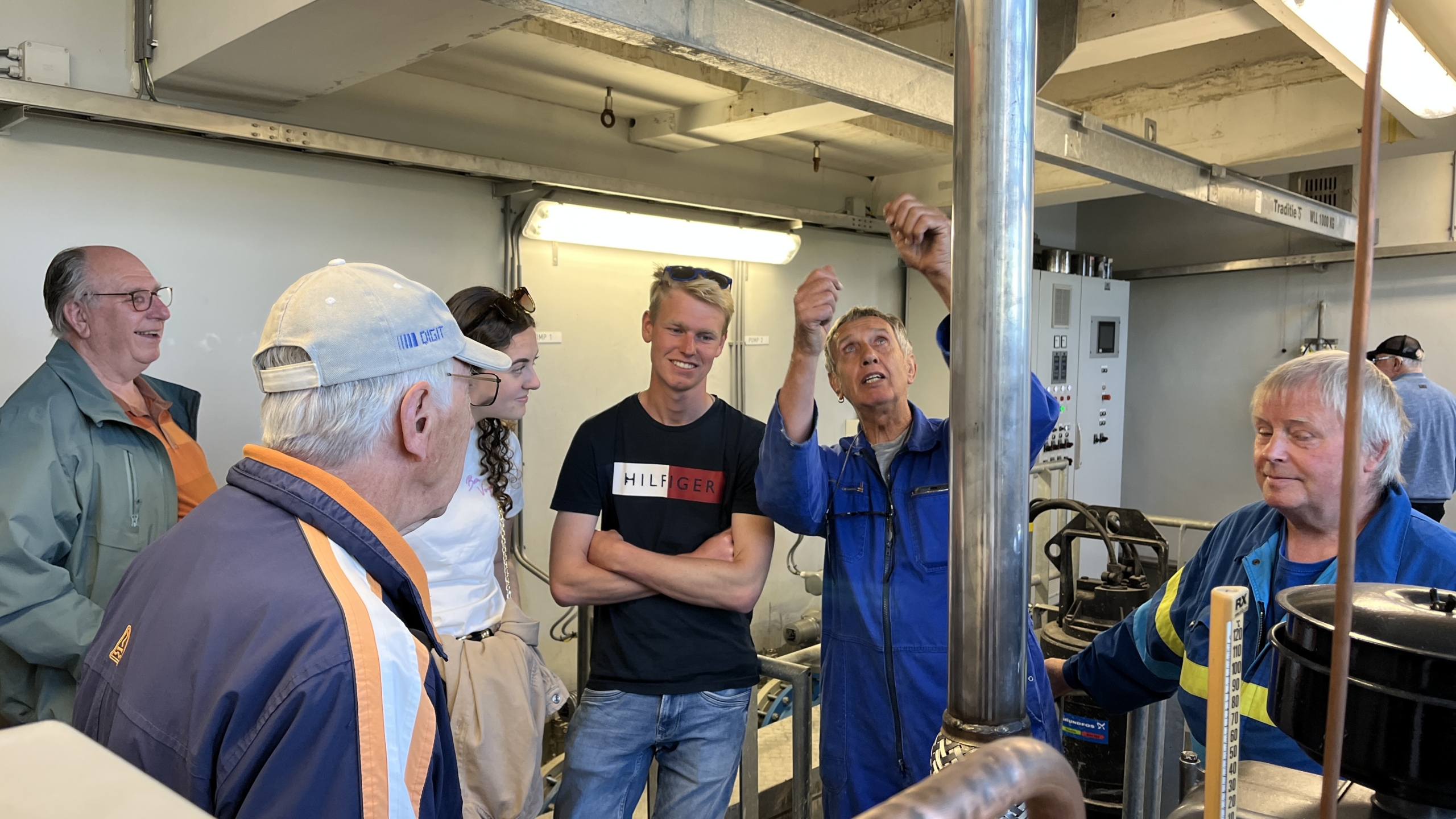 Een groep mensen luistert naar een man in blauwe overall die iets uitlegt in een industriële omgeving met metalen buizen.