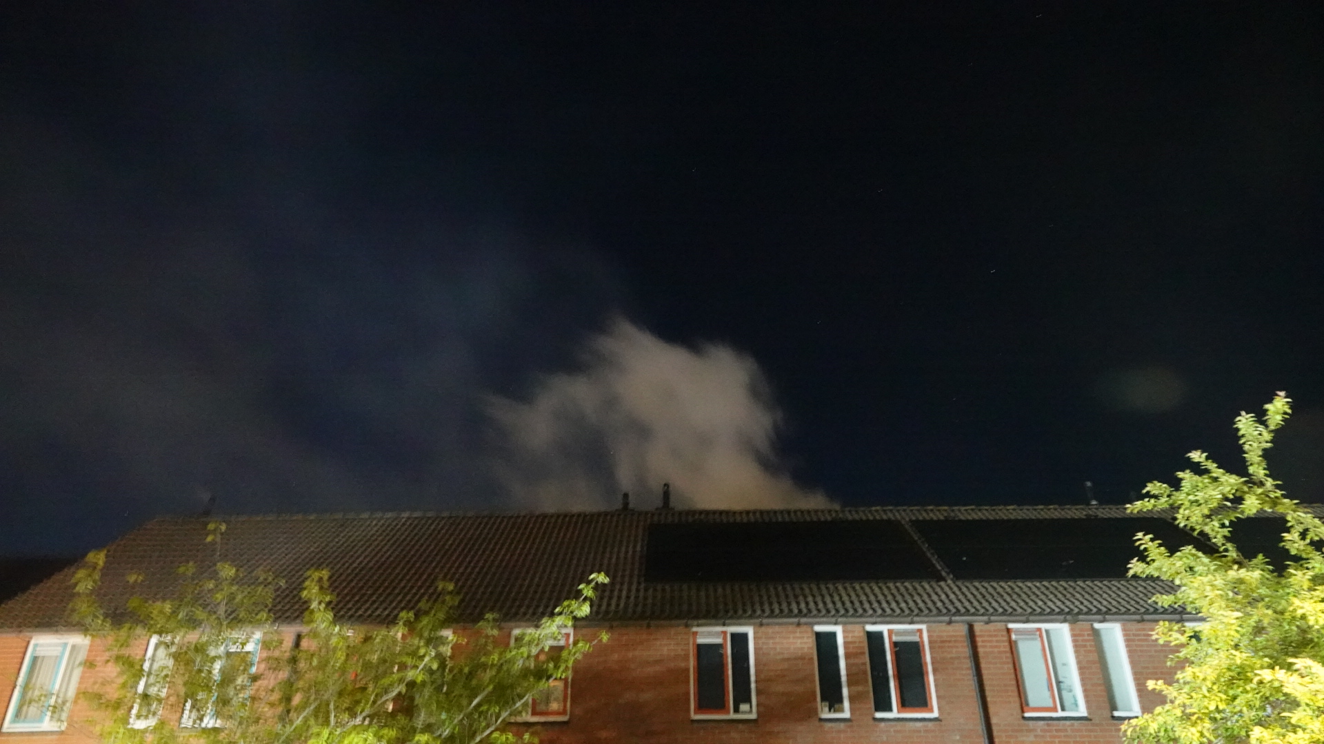 Rook stijgt op boven het dak van een bakstenen huis, gefotografeerd 's nachts met bomen op de voorgrond en een donkere lucht op de achtergrond.
