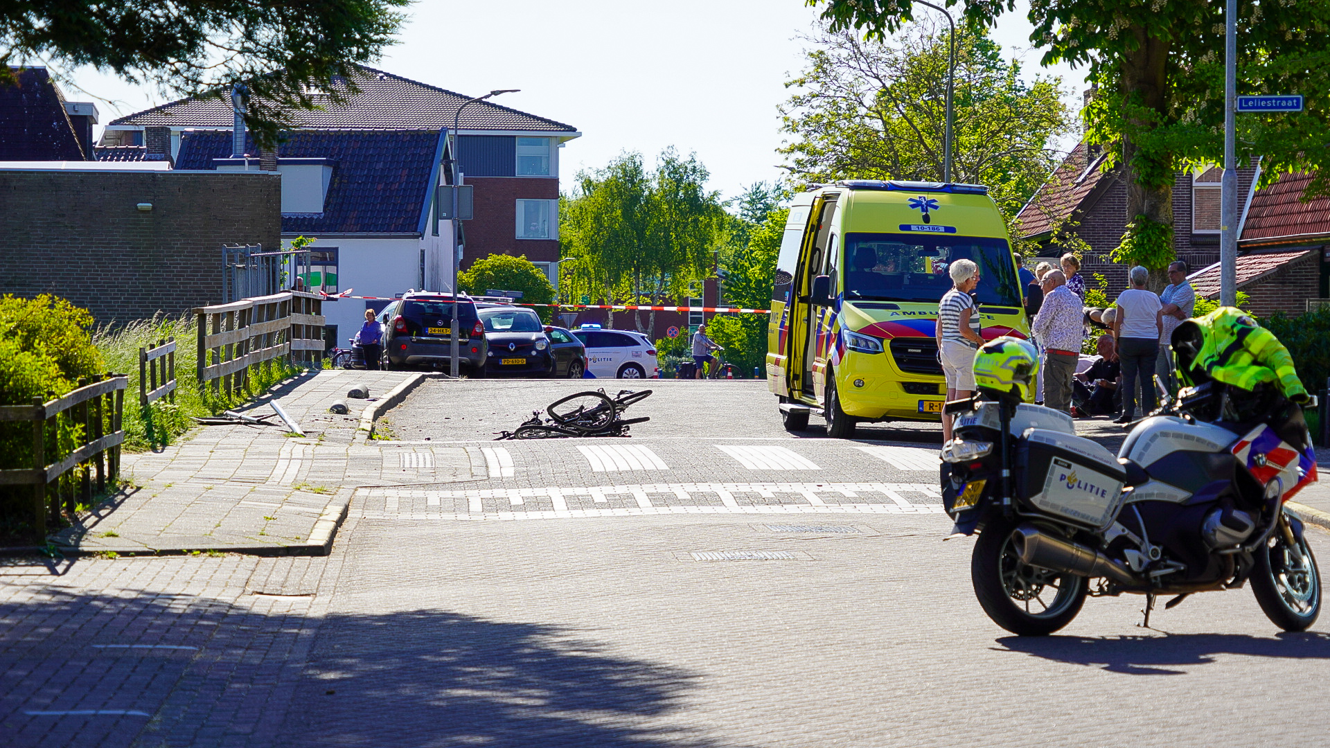 Een verkeersongeval waarbij een fiets op de straat ligt bij een zebrapad, omringd door een ambulance en politievoertuigen. Omstanders staan in de buurt, terwijl een politieagent bij een motorfiets staat. De omgeving is een woonwijk met bomen en huizen.