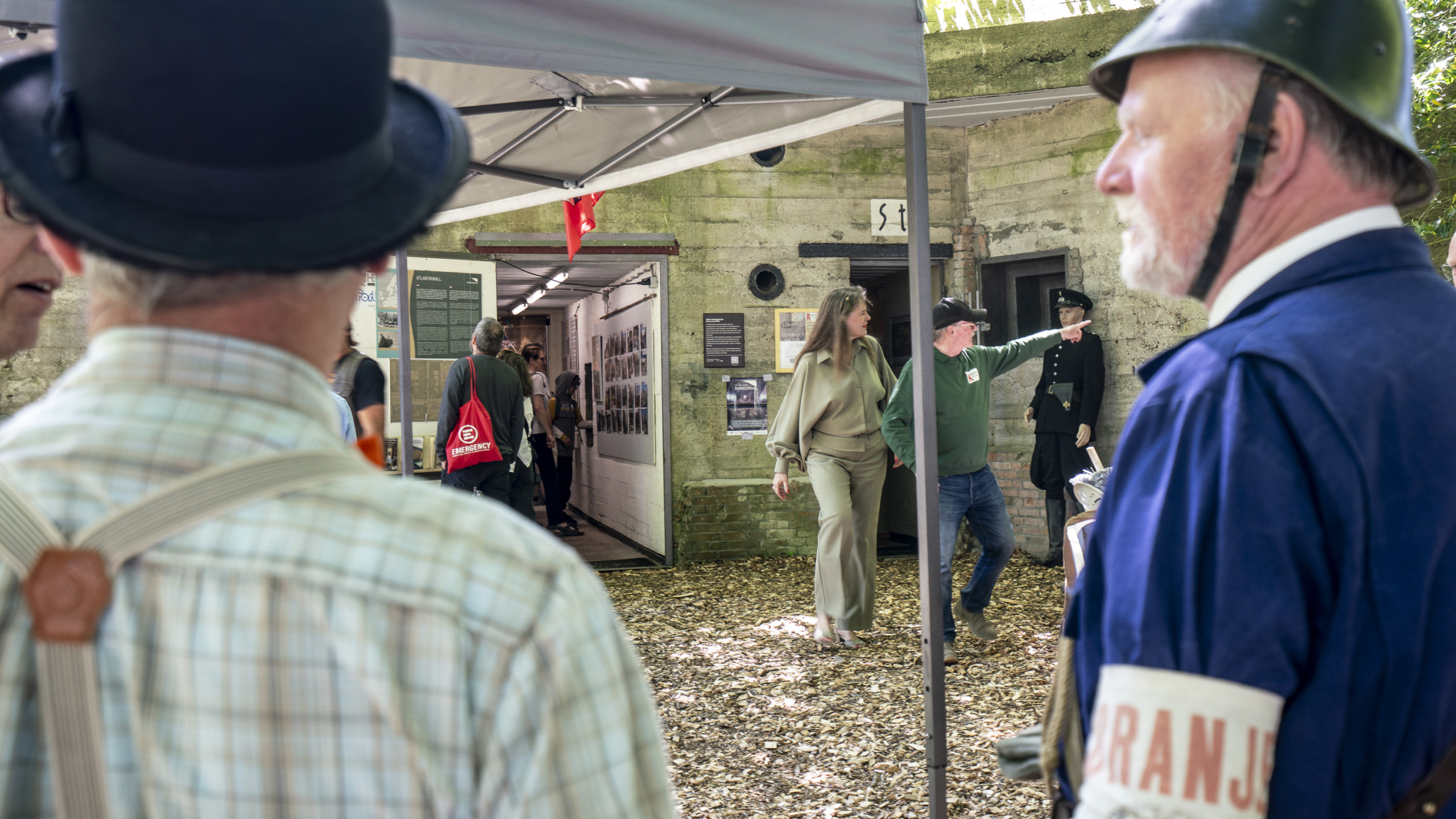 Mensen bij een open bunker, deels bedekt door een tentdoek; sommigen dragen kostuums uit de Tweede Wereldoorlog, terwijl anderen de ruimte binnen verkennen; een man wijst naar een mannequin in uniform.