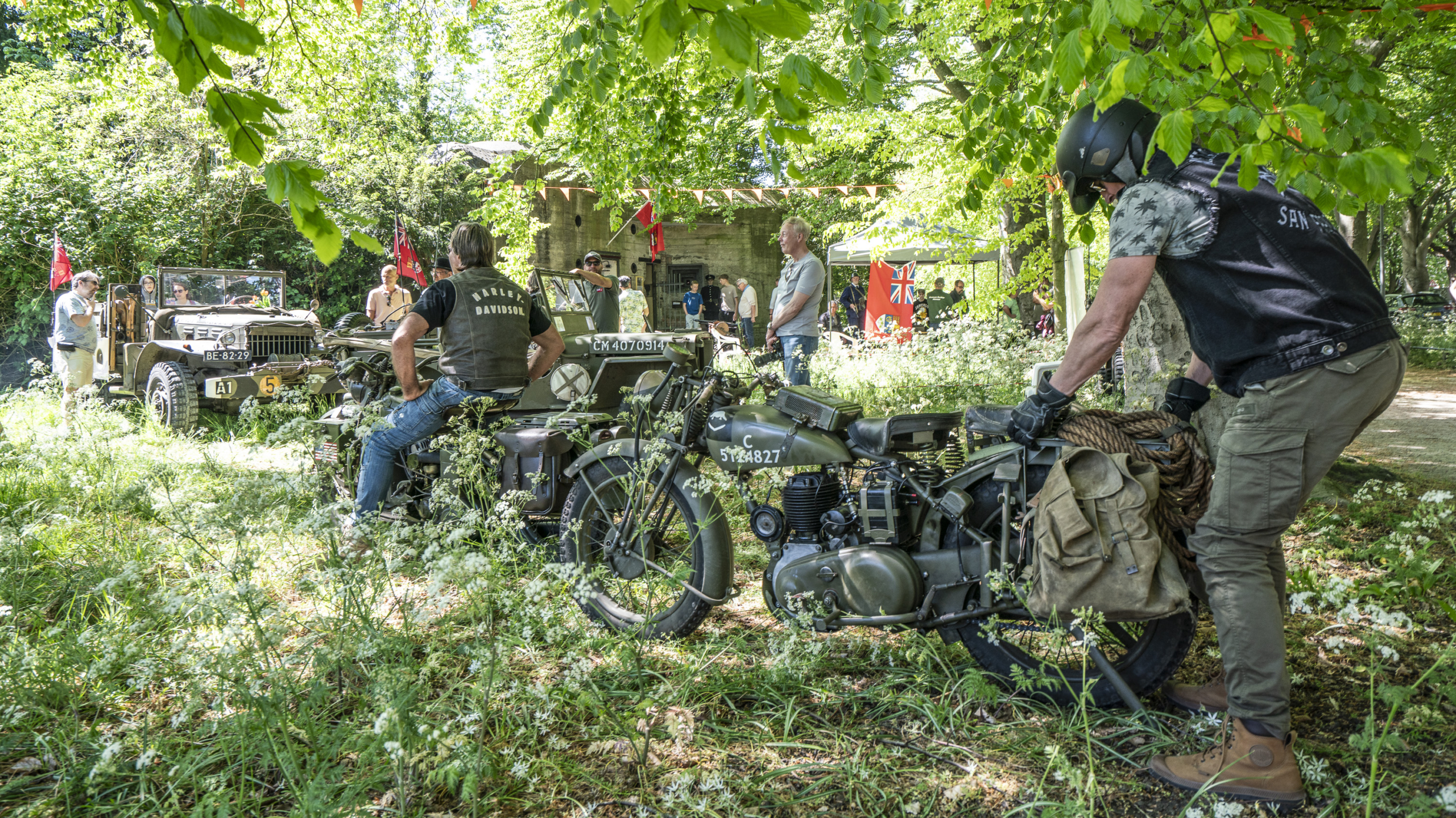 Mensen verzamelen zich rond oude miltaire voertuigen en motorfietsen in een groene open plek met veel bomen en gras. Er zijn een paar vlaggen en een tent met toeschouwers op de achtergrond.