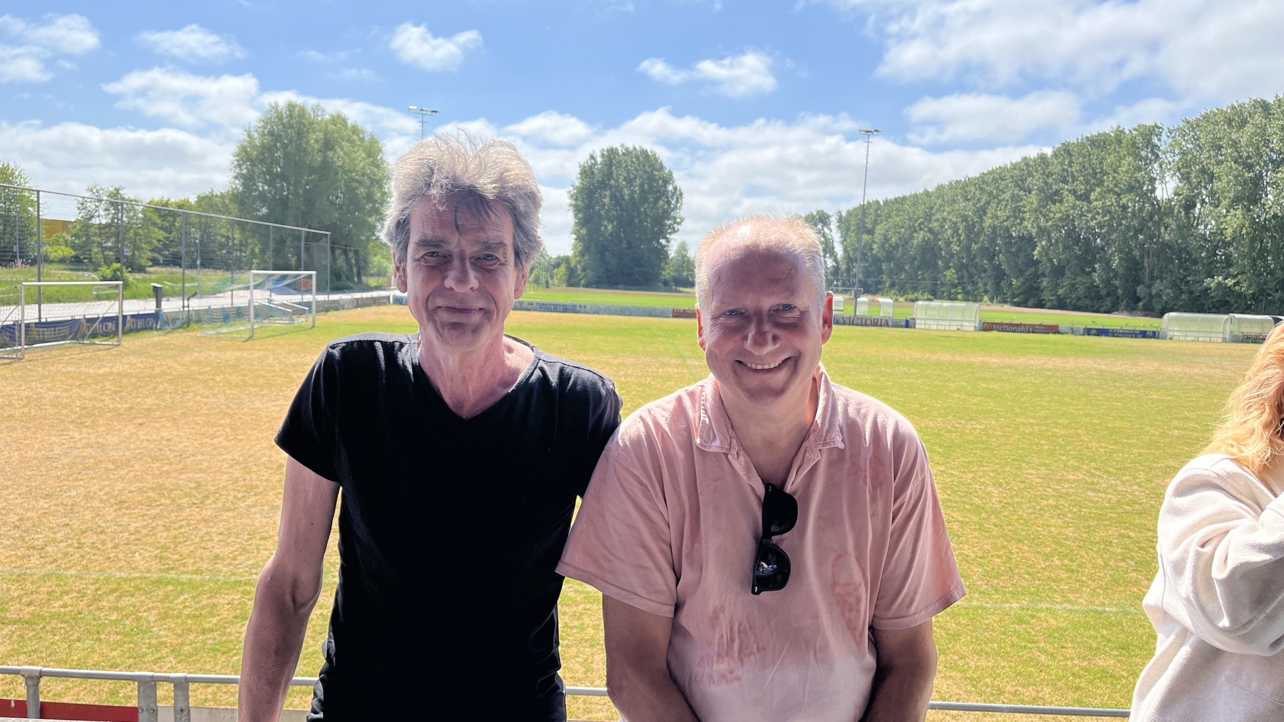 Twee mannen staan naast elkaar op een zonnig sportveld, met een heldere blauwe lucht en groene bomen op de achtergrond, en dragen respectievelijk een zwart en een lichtroze shirt.
