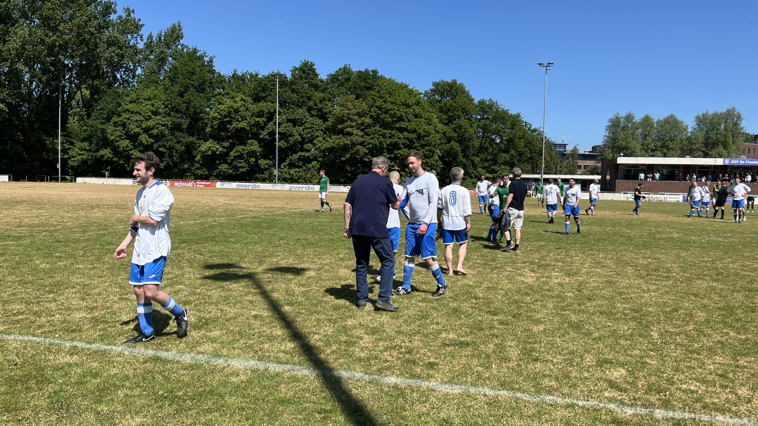 Voetbalspelers en toeschouwers verzamelen zich op een zonnig sportveld na de wedstrijd, met een achtergrond van bomen en een tribune.