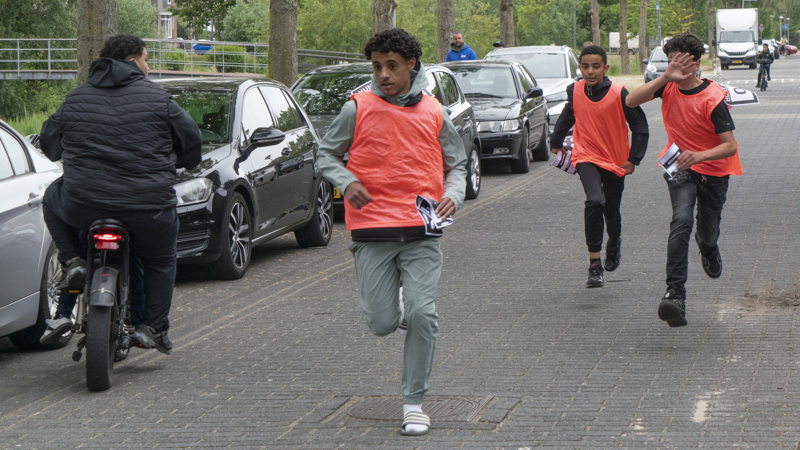 Groep jongeren, gekleed in oranje hesjes, rent over een trottoir naast geparkeerde auto's, terwijl een persoon op een scooter in tegenovergestelde richting rijdt.