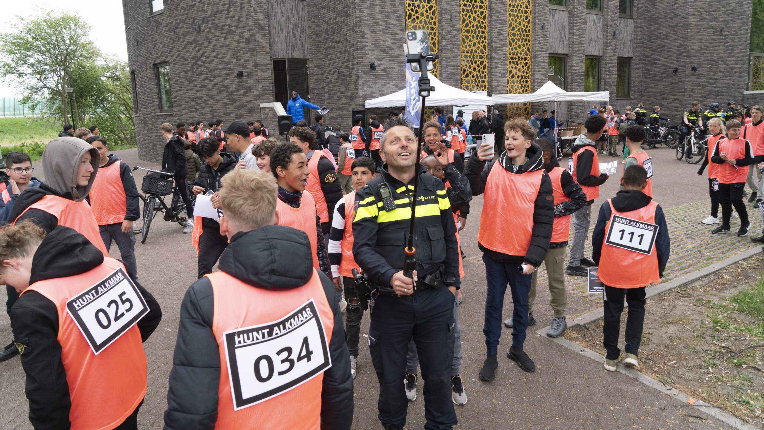Groep jongeren in oranje hesjes met nummerborden verzamelt zich buiten bij een gebouw. Een agent maakt een selfie met een selfiestick, terwijl anderen op de achtergrond staan met fietsen en politiebewaking zichtbaar is. Er staan tenten en mensen in de buurt van de ingang van het gebouw.