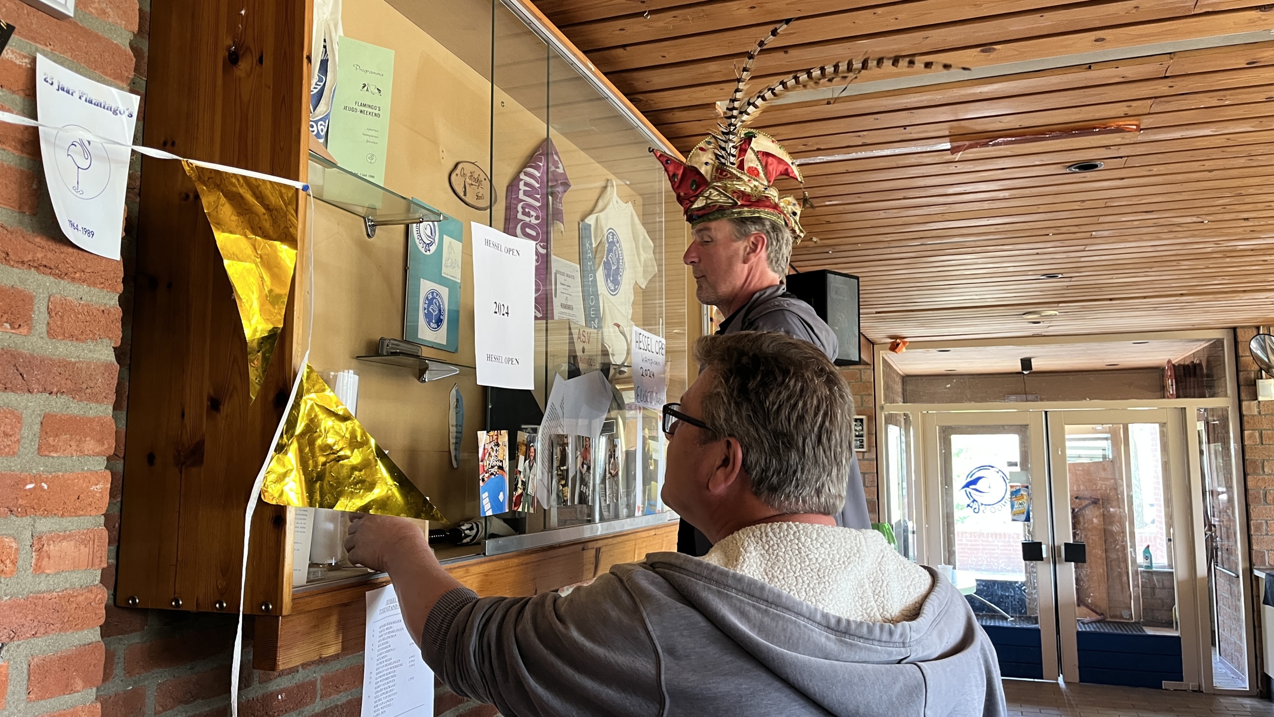 Twee mannen staan voor een glazen vitrine met documenten en souvenirs, waarbij de man rechts een opvallende, felgekleurde hoed met veren draagt. Er hangen gouden vlaggetjes en papier aan de linkerkant van de kast. De ruimte heeft een houten plafond en bakstenen muren.
