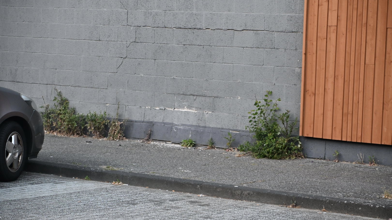 Een grijze stenen muur met scheuren, een geparkeerde auto aan de linkerkant, en enkele plantjes die langs de stoep groeien.
