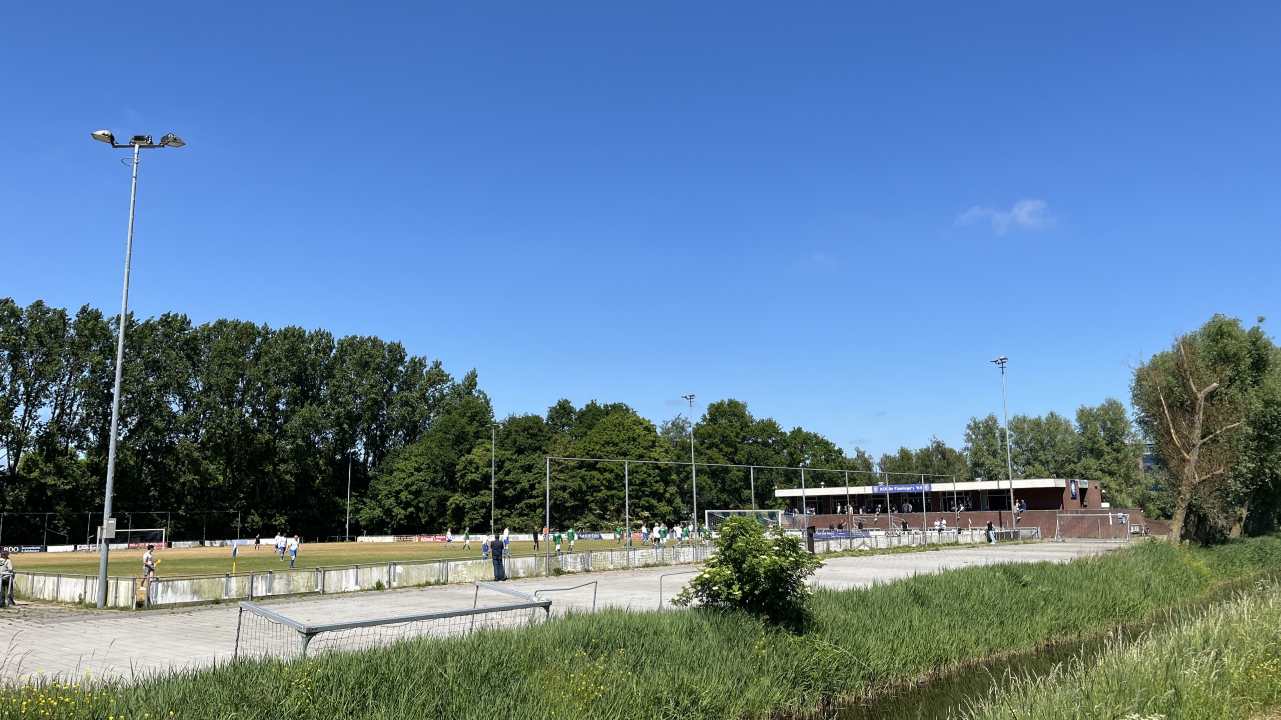 Voetbalveld met spelers en toeschouwers aan de rand, omgeven door bomen en groen gras.