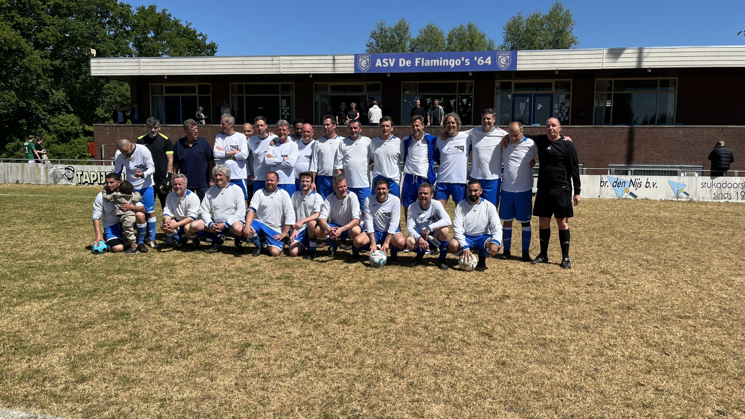 Een groep voetballers poseert voor een teamfoto op een grasveld, gekleed in witte en blauwe tenues, met een bord van ASV De Flamingo's '64 op de achtergrond.