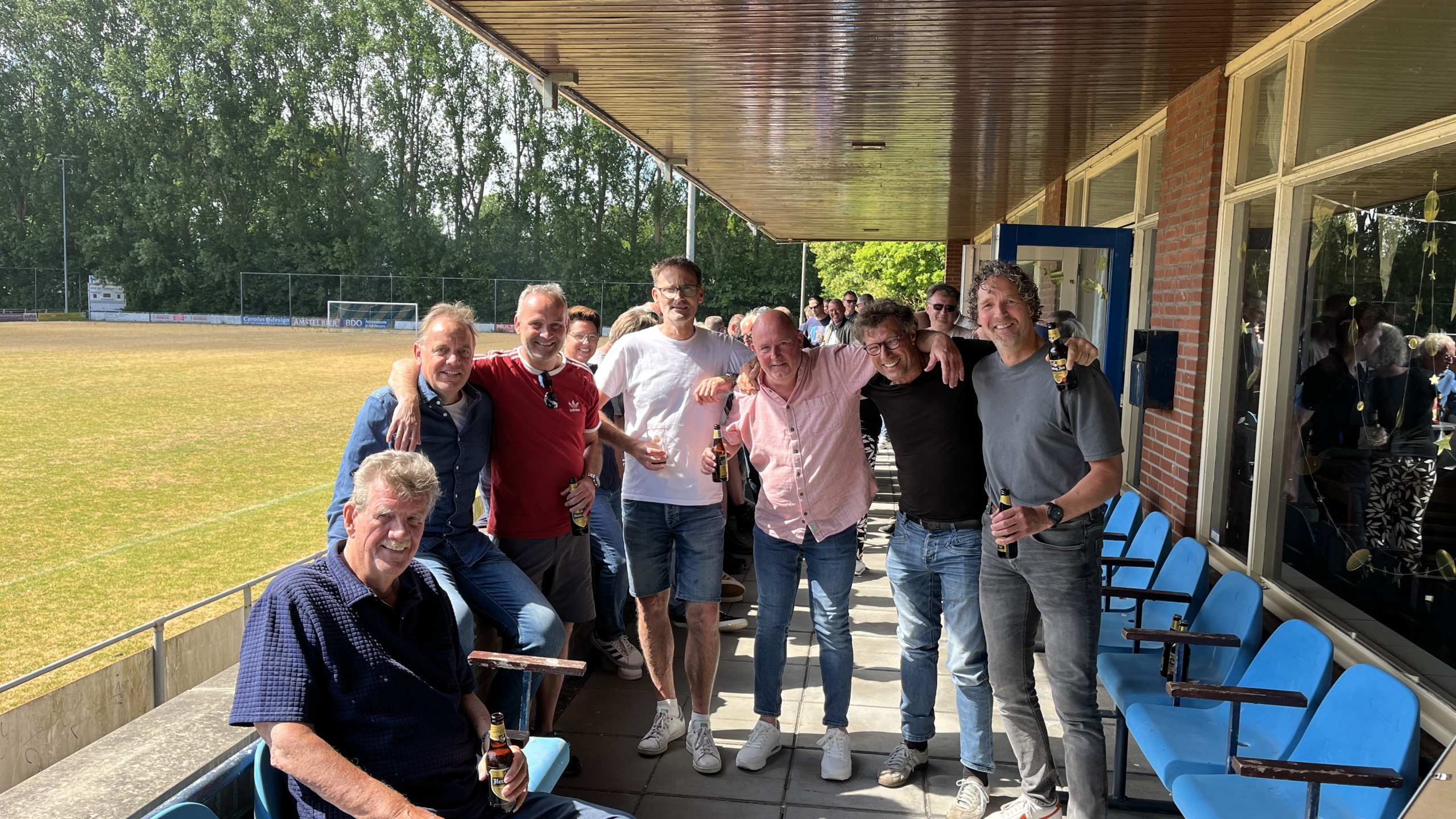 Een groep mannen poseert lachend voor een sportveld, met enkele stoelen op de voorgrond en een glazen wand rechts; sommigen houden drankflesjes vast.