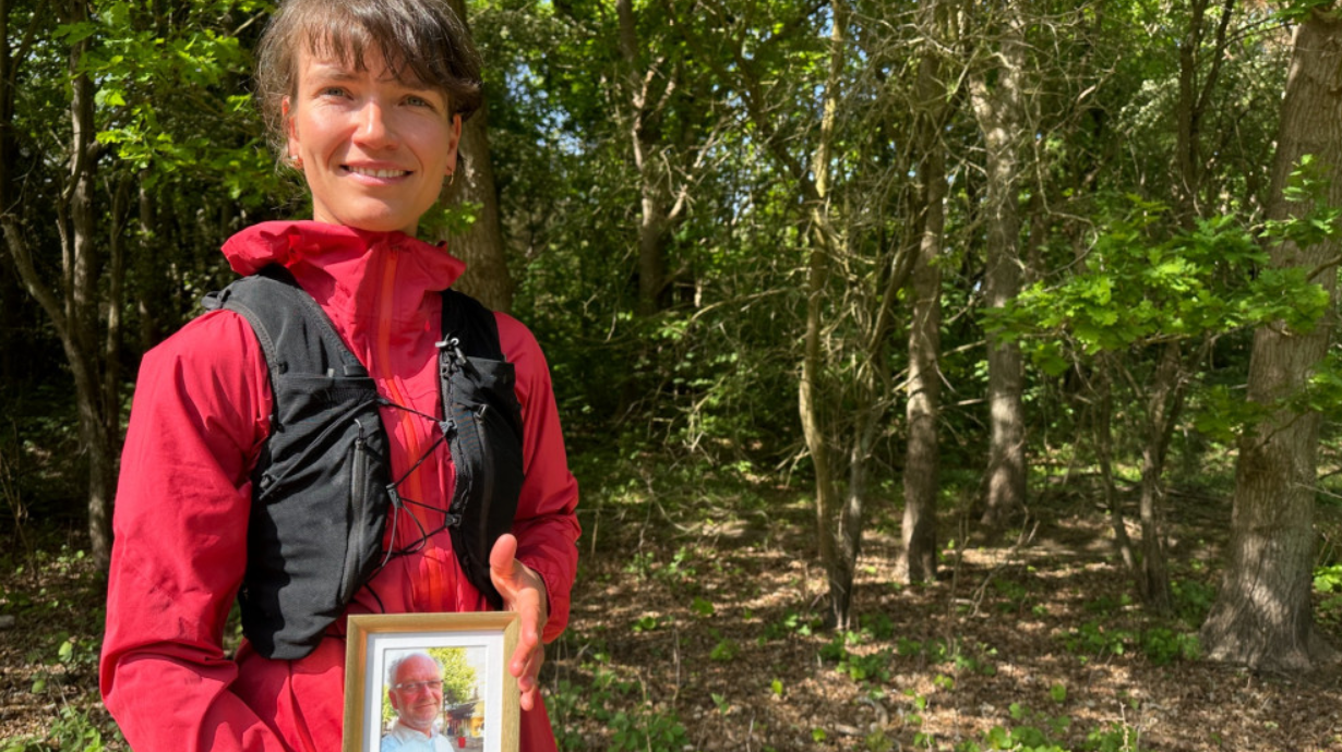 Vrouw in een rood jack staat buiten voor een bos en houdt een ingelijste foto van een oudere man vast.