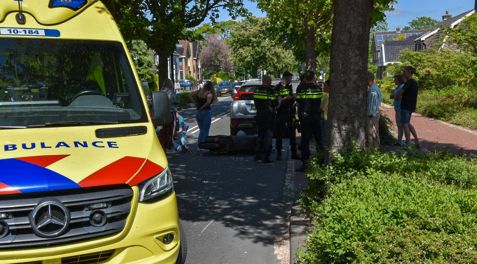 Geel ambulancevoertuig staat geparkeerd naast een weg, omringd door politieagenten en omstanders. Een scootmobiel ligt op de grond in de buurt van een boom, terwijl een persoon knielt en een andere persoon toespreekt. Het is een zonnige dag met groene bomen langs de straat.