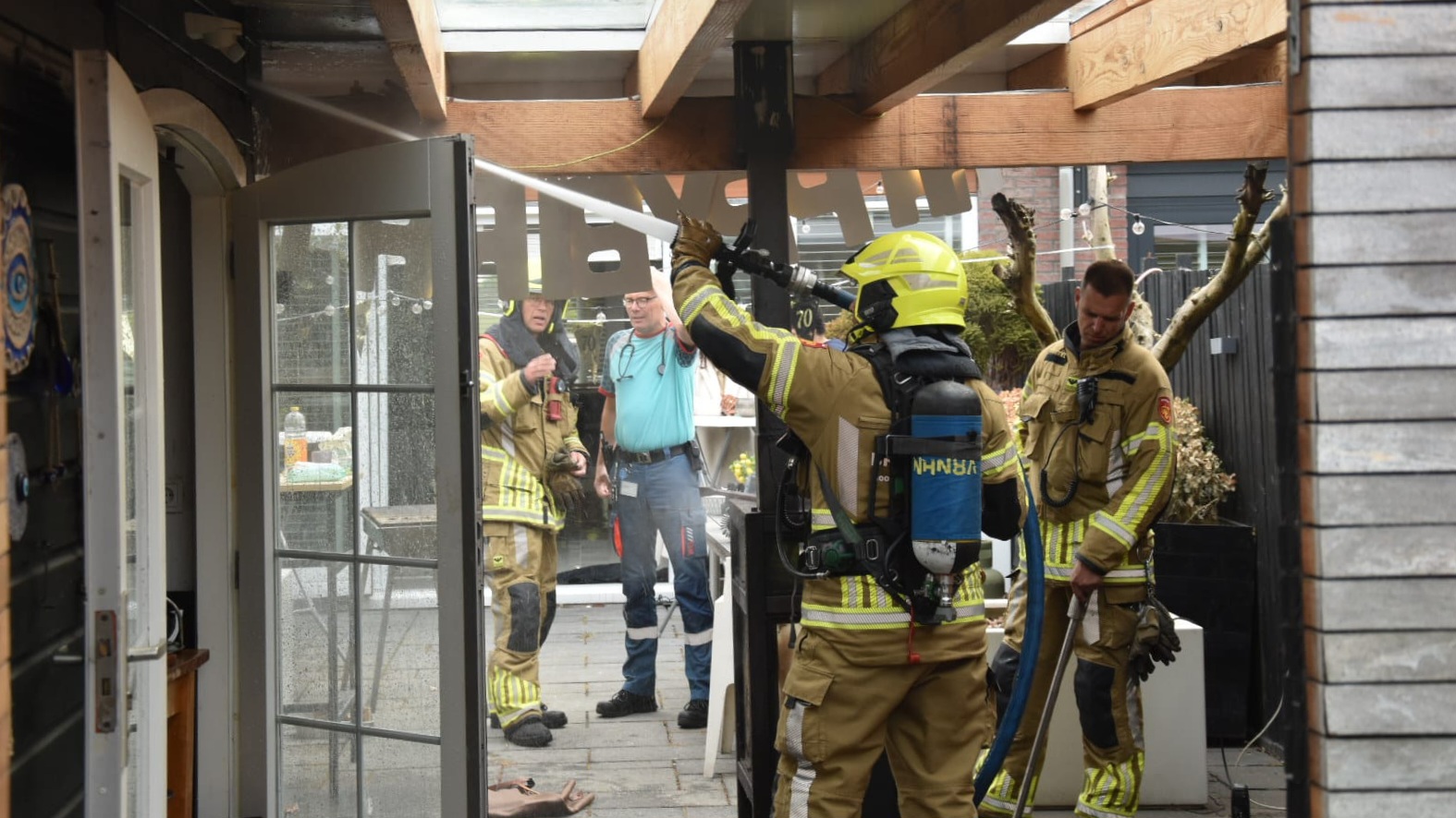 Brandweerlieden met beschermende kleding blussen een brand in een overdekte patio, terwijl andere personen toekijken en helpen.