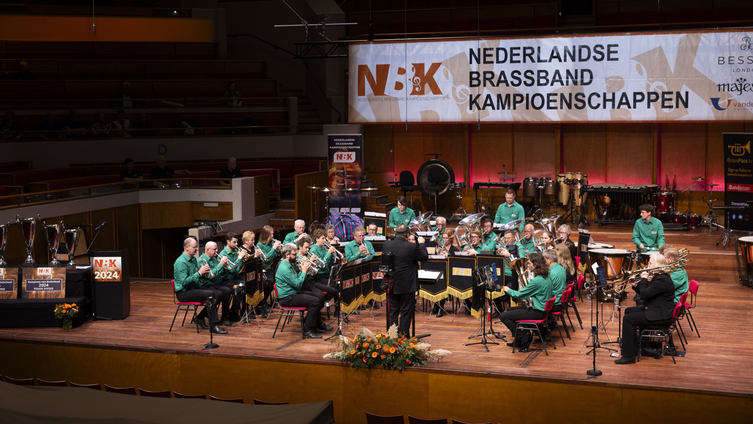 Mensen in groene uniformen spelen blaasinstrumenten tijdens de Nederlandse Brassband Kampioenschappen, met een dirigent aan de voorkant en een groot bord met de tekst 'Nederlandse Brassband Kampioenschappen' op de achtergrond. Trofeeën staan links op het podium.