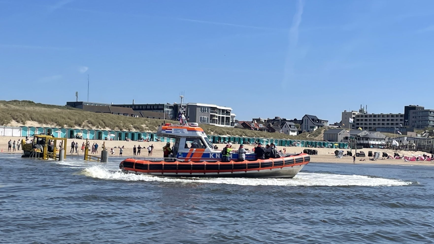 Reddingsboot op zee voor een drukbezocht strand met badgasten en strandpaviljoens onder een heldere blauwe lucht.