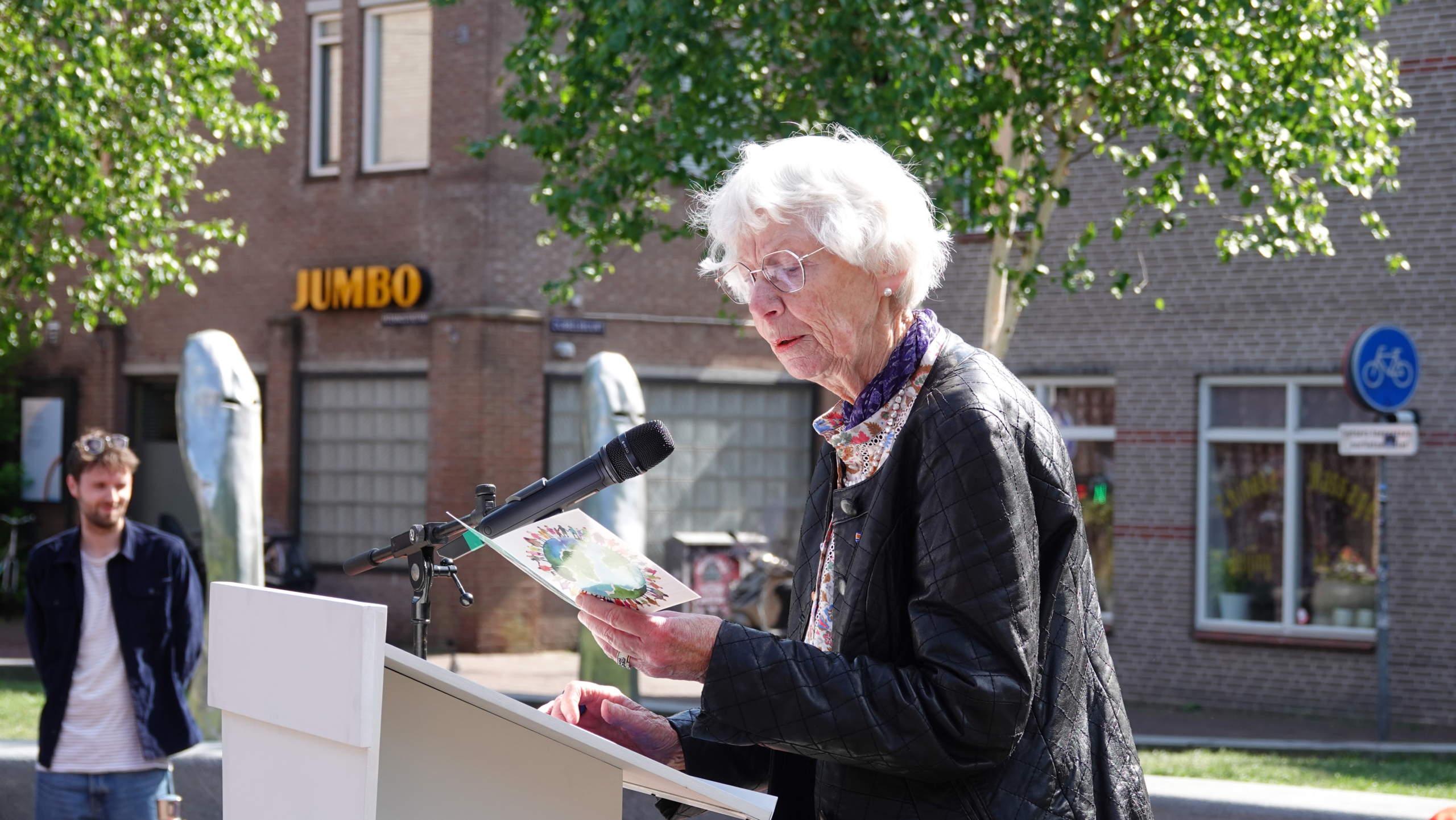 Een oudere vrouw leest voor bij een microfoon op een open locatie, met een jongen op de achtergrond en een gebouw met "JUMBO" zichtbaar.